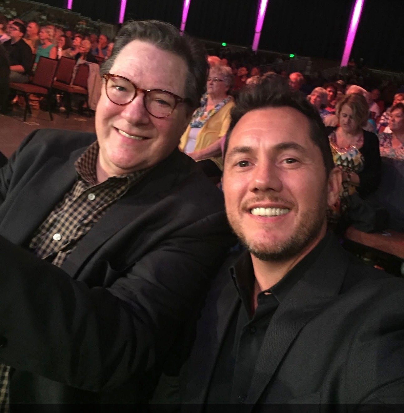Two smiling men taking a selfie at an event with a large audience seated behind them.
