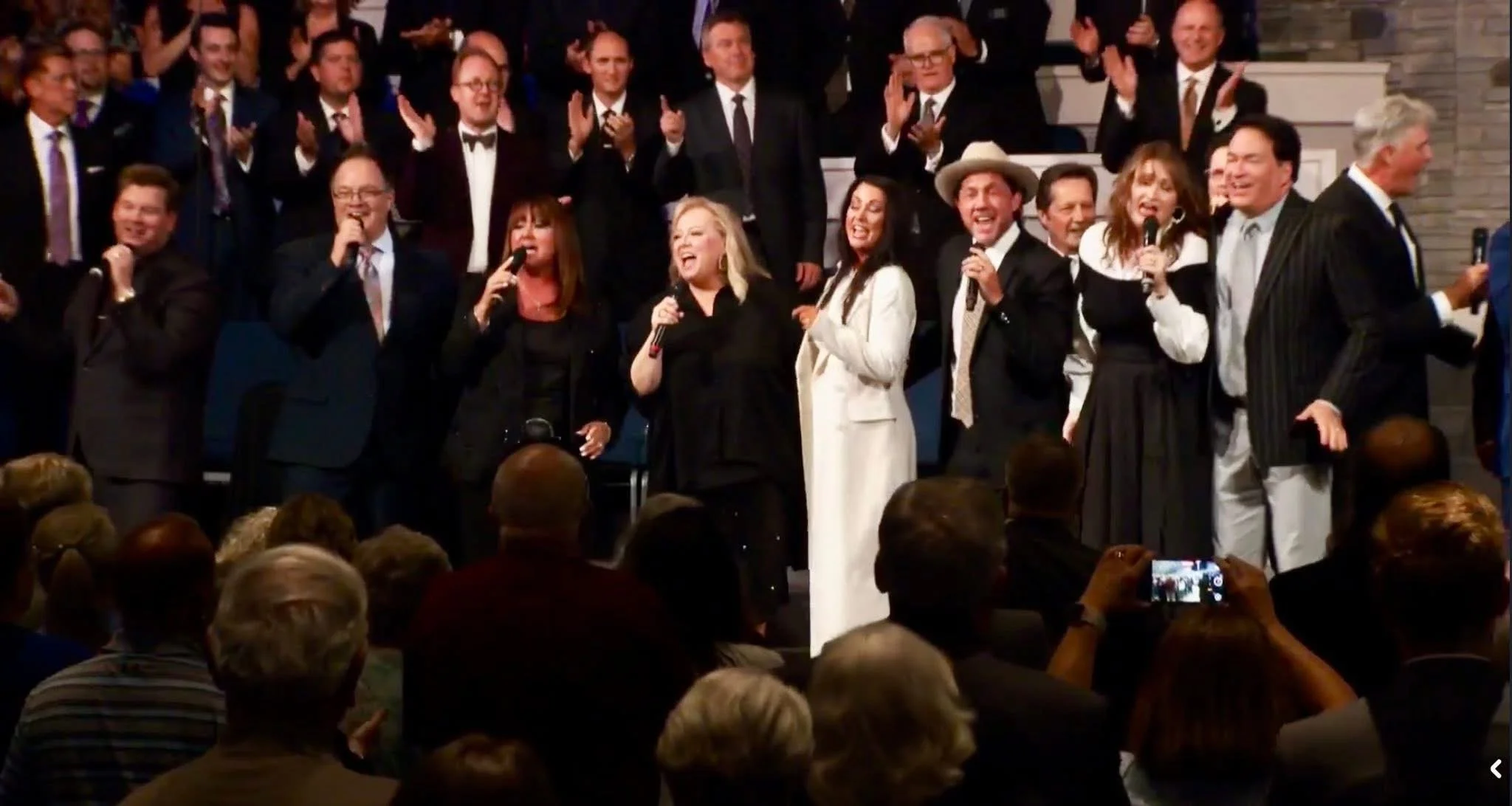 Group of people singing and celebrating on a stage, some holding microphones, with an audience watching.