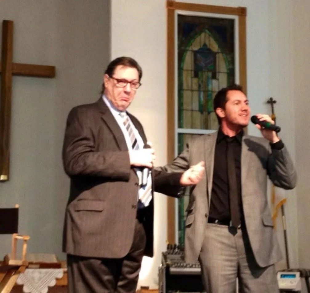 Two men in suits stand on a stage, one holding a microphone and the other making a gesture with his hand, inside a church with stained glass windows in the background.