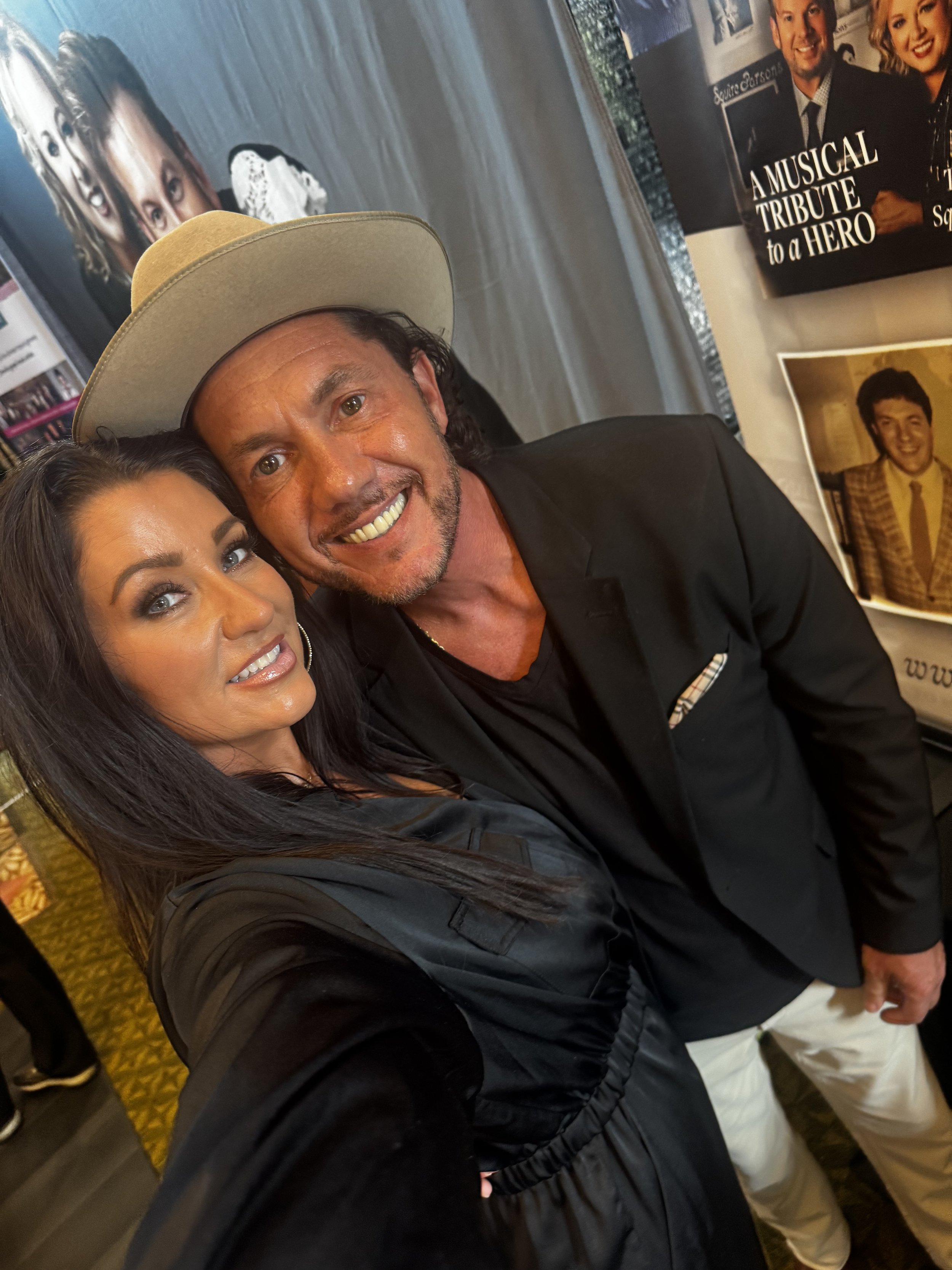A woman and a man take a selfie together at an event, with posters and photographs in the background that include a musical tribute to a hero and a person in a plaid suit.