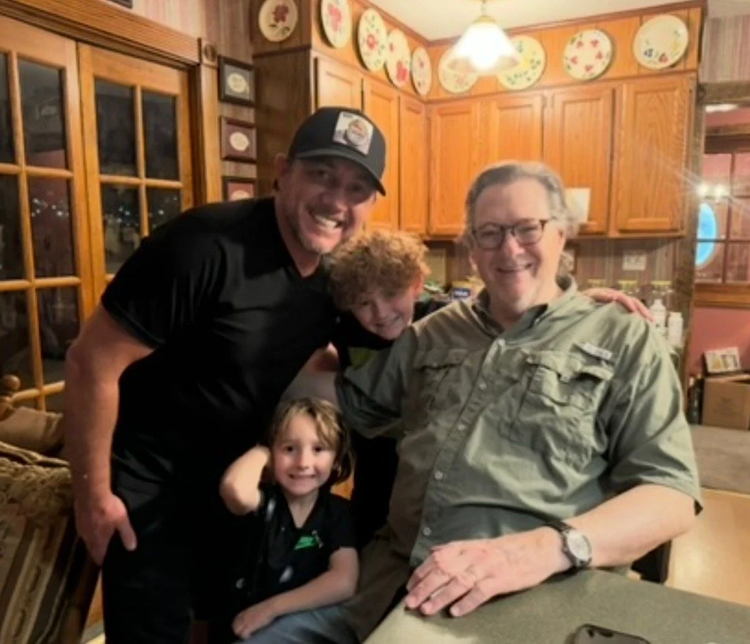 Four people smiling and posing together inside a warmly lit kitchen, with wooden cabinets and decorative plates on the wall.