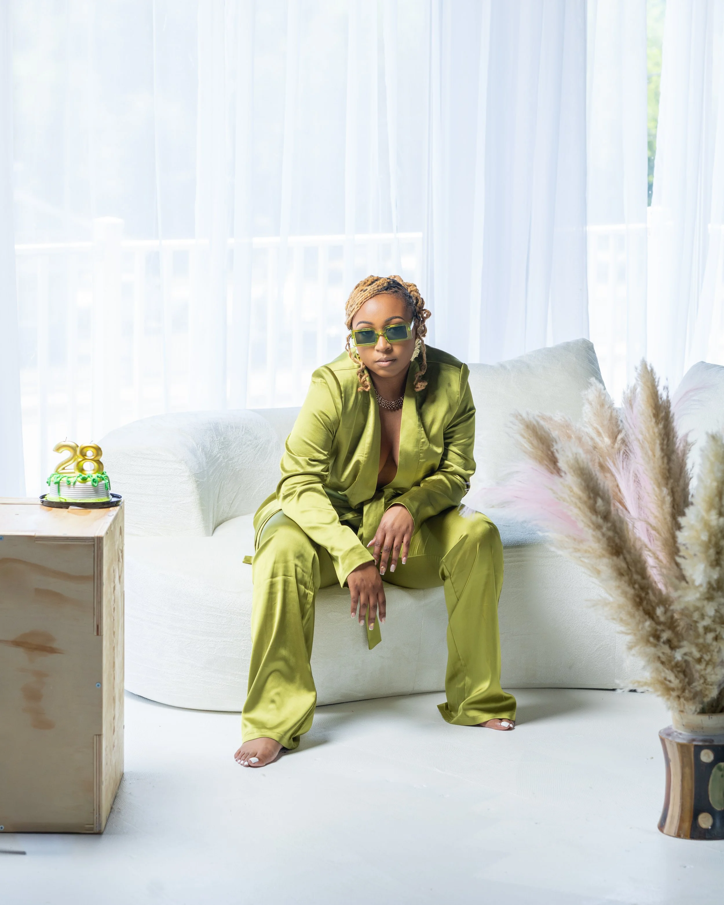 A woman wearing green satin pajamas and sunglasses sitting on a white couch in a bright room with sheer curtains, a birthday cake with the number 28 on top, and pampas grass in a vase.