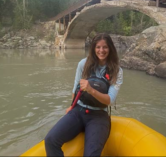 Woman smiling and sitting in a yellow inflatable boat on a river, holding a paddle, with a stone bridge and trees in the background, Professional guided rapid rafting tours at Idaho Springs Rafting Company, Colorado