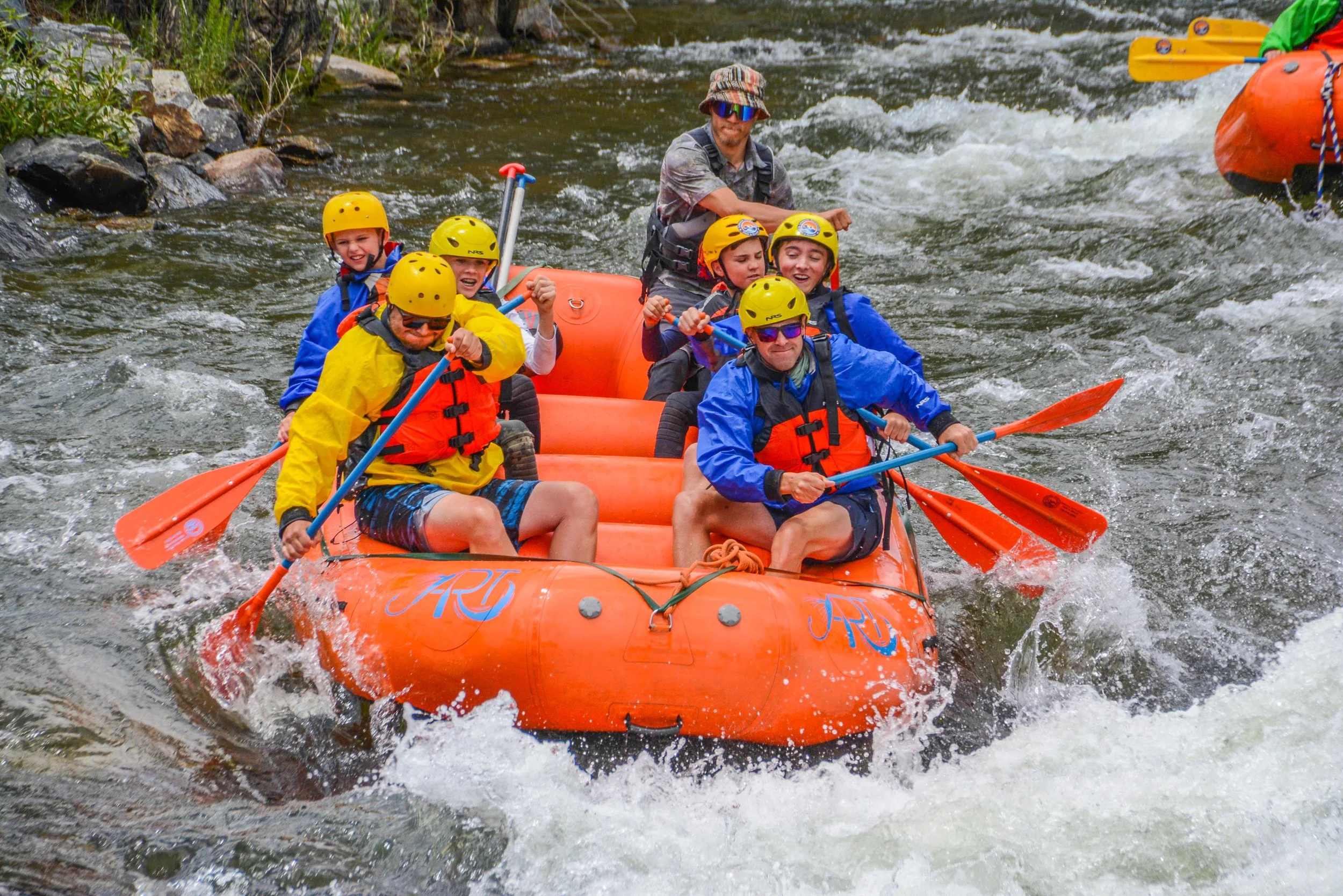 A group of six children and one adult rafting on a river in an orange inflatable boat. They are wearing helmets, life jackets, and paddling with orange oars.