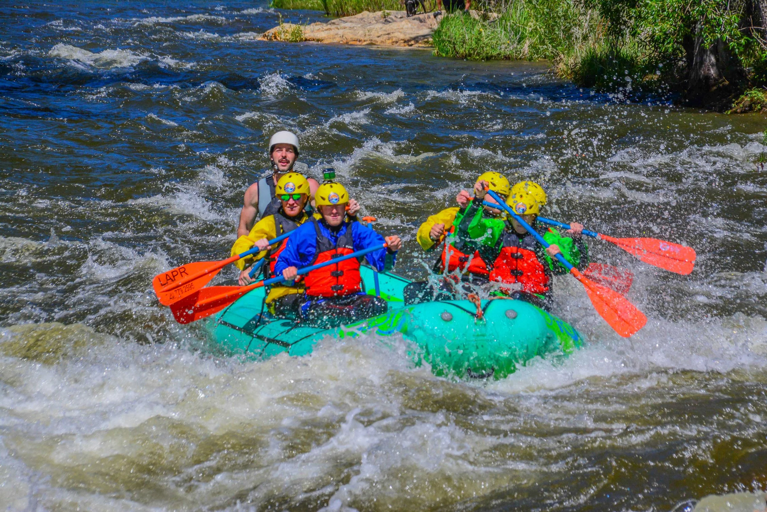 A group of people rafting on a river in a bright blue inflatable raft, wearing yellow helmets and life jackets, navigating through rushing water with paddles.