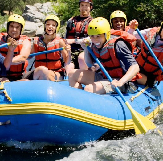 Group of six people in a blue inflatable raft, white water rafting, wearing yellow helmets and life jackets, smiling and paddling on a river surrounded by trees and rocks.