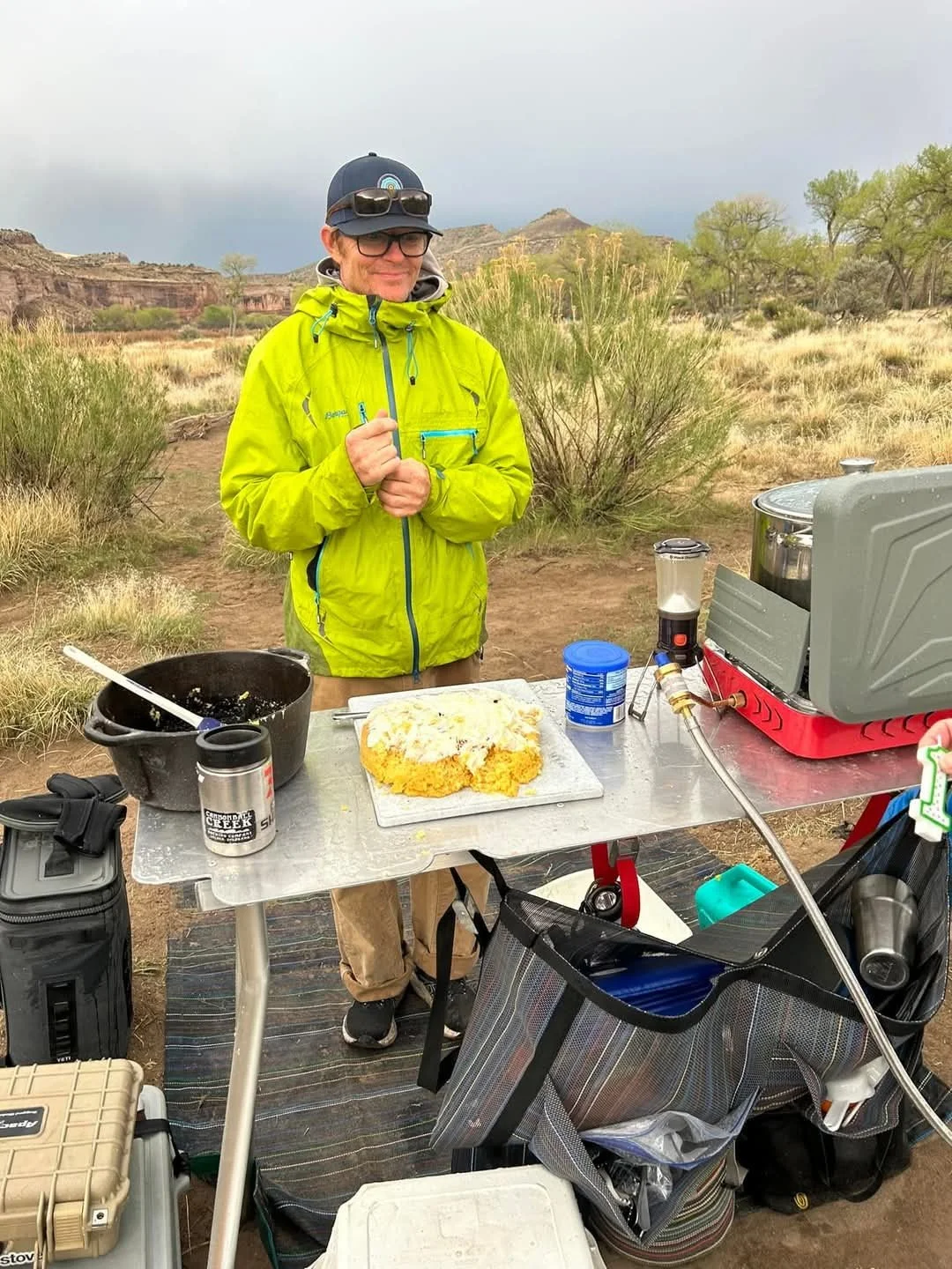 A woman in a bright yellow jacket and hat stands outdoors at a camping stove with food on a cutting board, surrounded by camping gear, in a desert-like landscape with sparse trees and rocky formations.