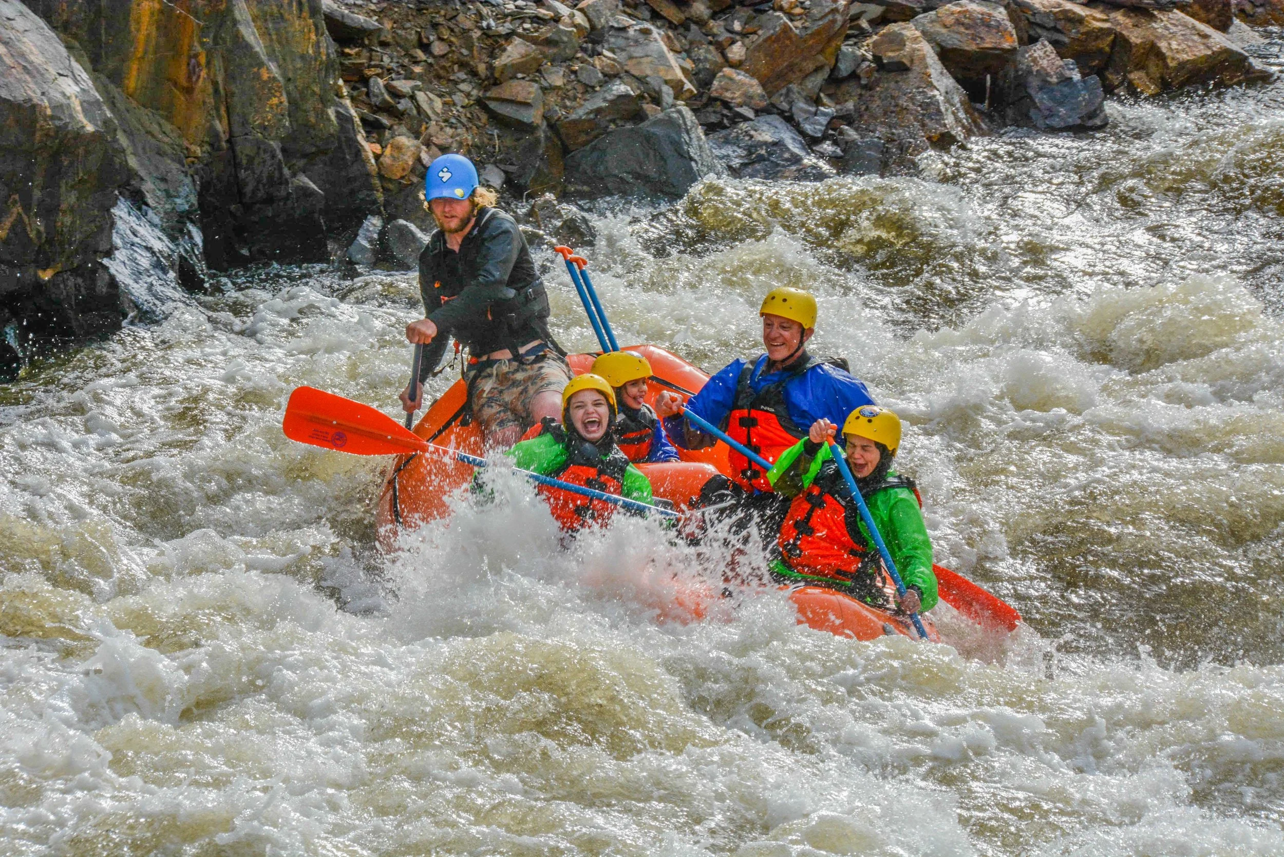 A group of people white water rafting in a river, wearing helmets and life jackets, with rocks and water splashing around.