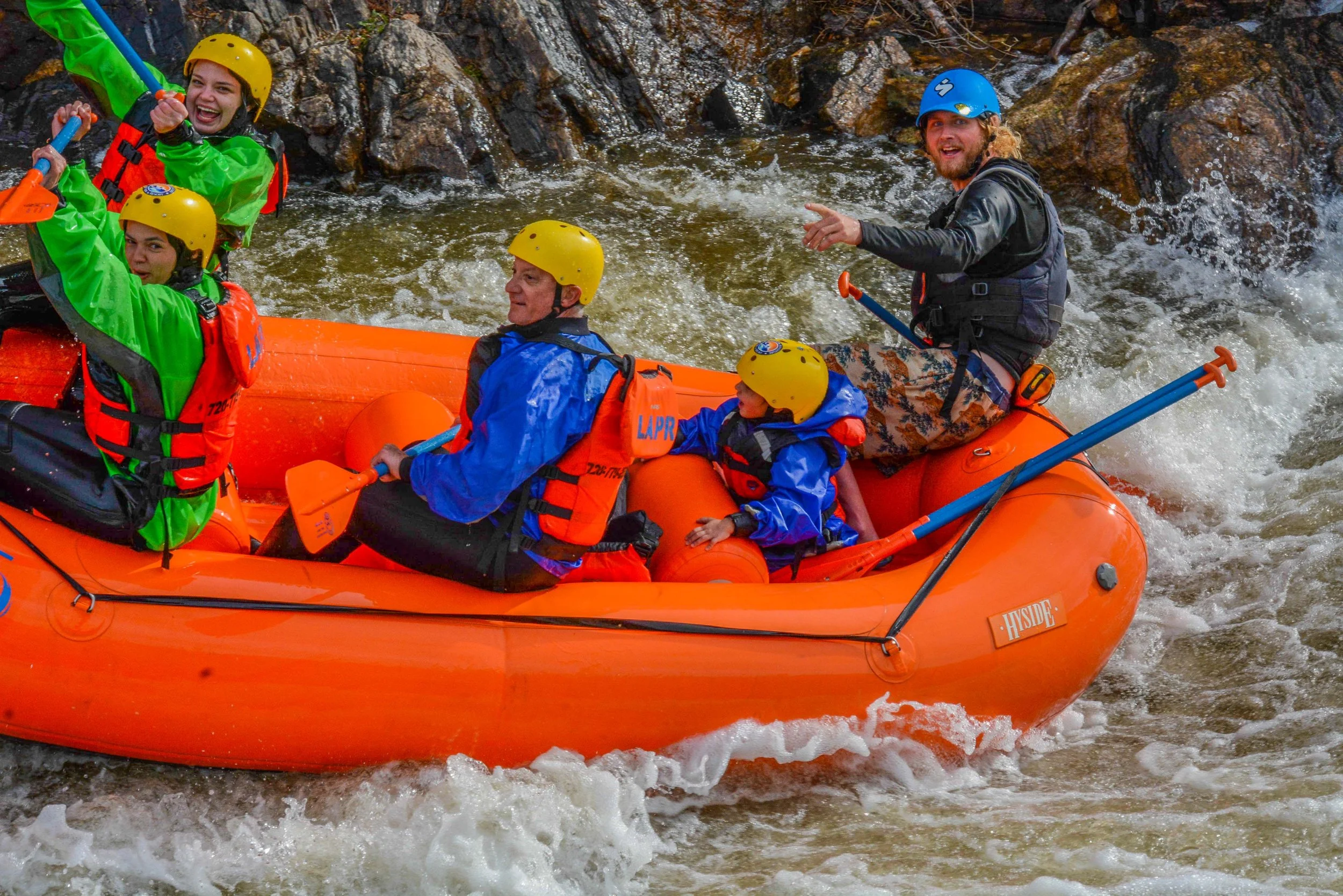A group of people, including children and adults, are rafting on a river in an orange inflatable raft. They are wearing life jackets and helmets for safety, and the surroundings are rocky with water splashing around.