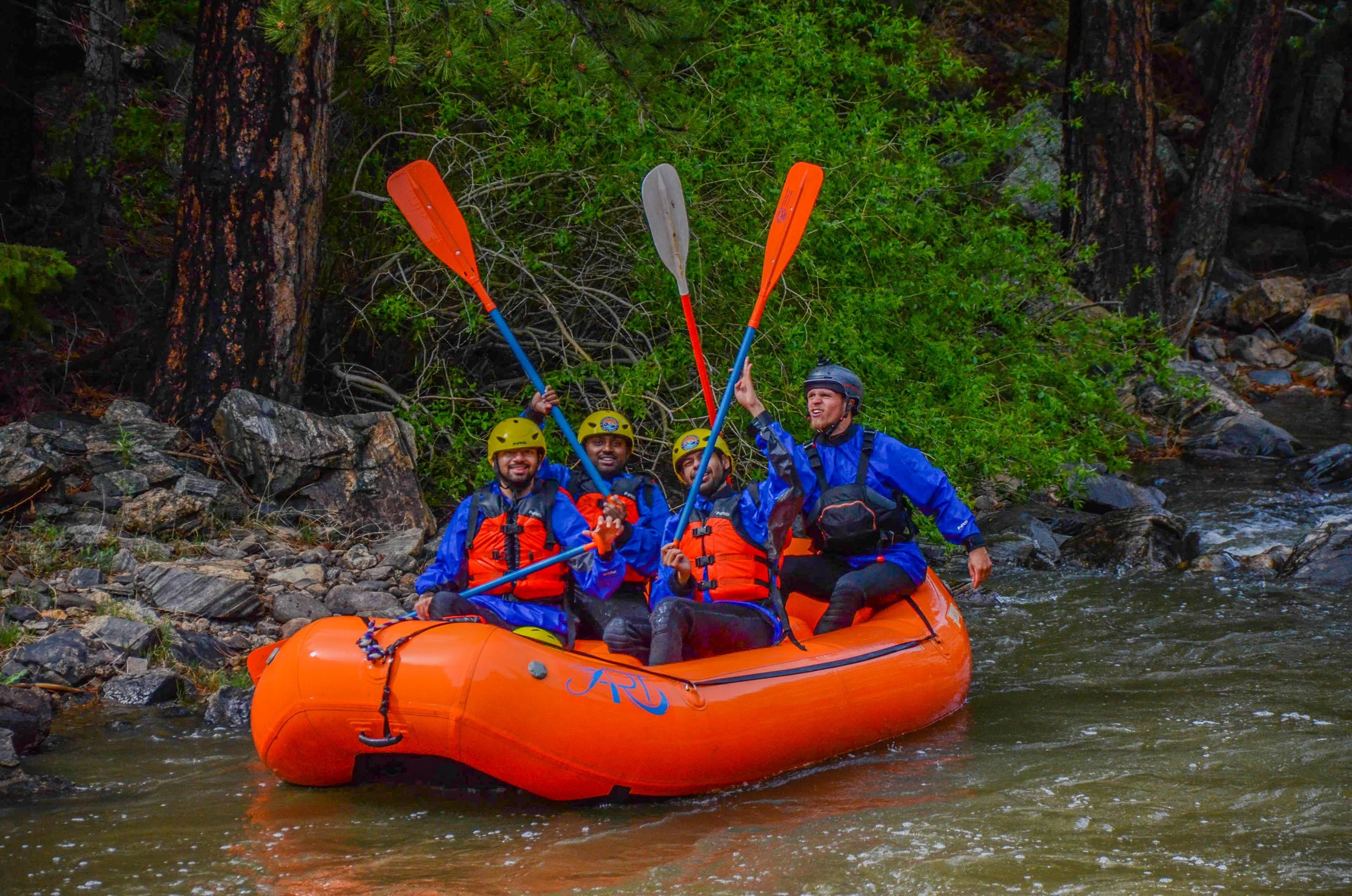 Four people in a bright orange inflatable raft on a river, surrounded by rocks and green trees, wearing helmets and life jackets, holding paddles.