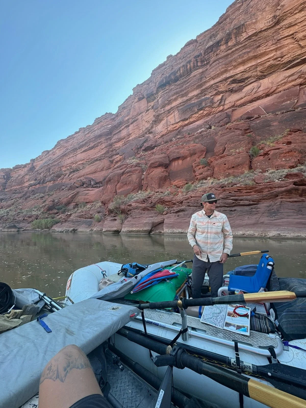 A man standing on a boat filled with outdoor gear, beside a river with red rock cliff walls in the background.