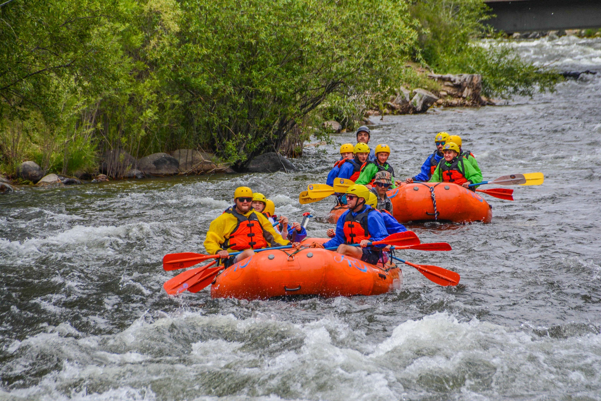 Group of people wearing helmets and life jackets rafting on a river surrounded by greenery.