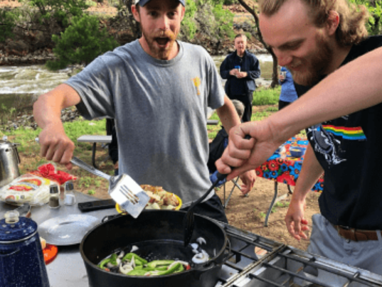 Two smiling men cooking on an outdoor stove, one stirring a pan with vegetables while the other holds a spatula, with a group of people in the background near a river and trees.