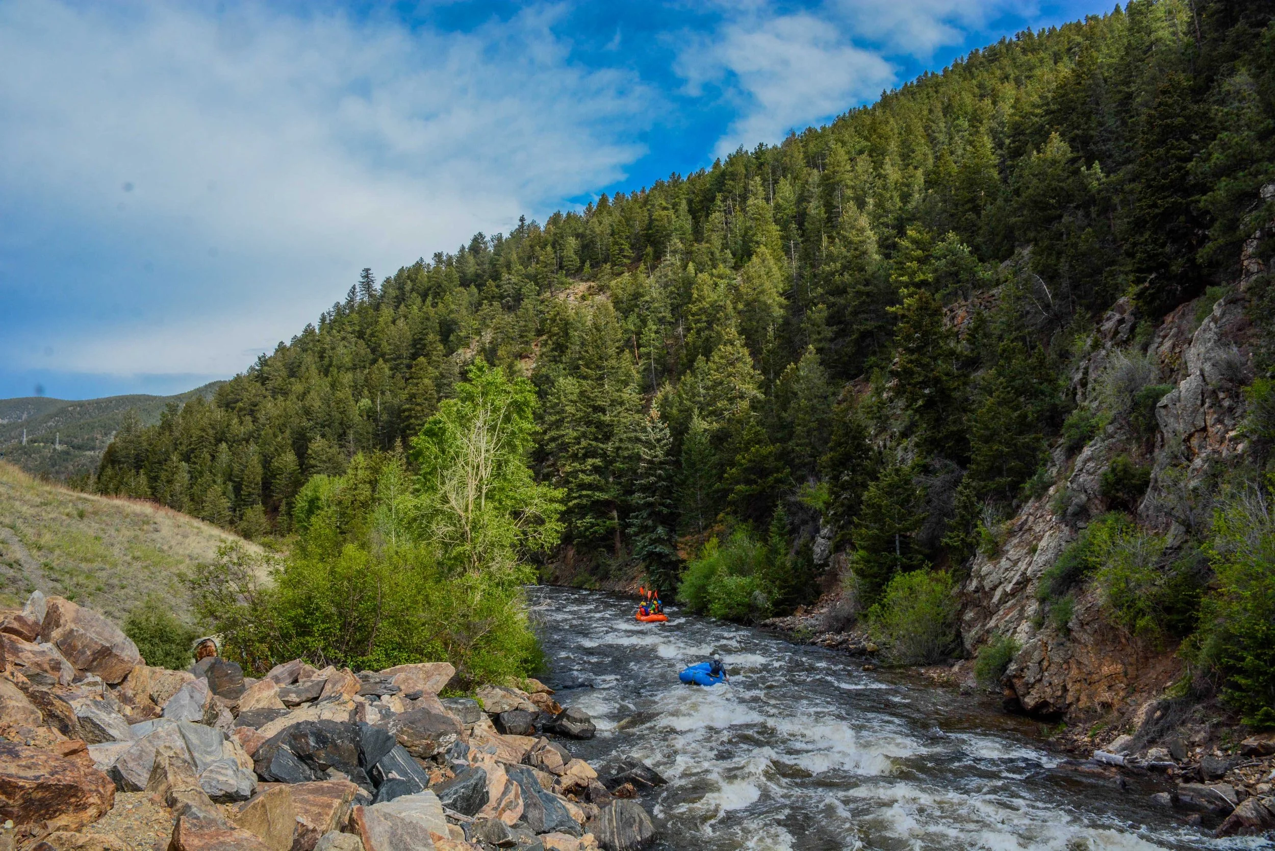 Two people in inflatable rafts white water rafting on a river surrounded by green forest on a cloudy day.