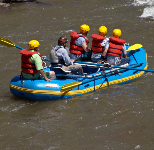 Group of six people rafting down a river in an inflatable blue raft, all wearing yellow helmets and red life jackets.