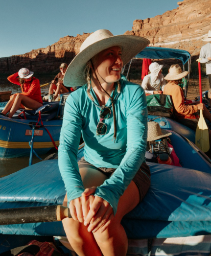Woman wearing a wide-brimmed hat and sunglasses sitting on an inflatable boat in a river, surrounded by other people in hats and casual clothing, with canyon cliffs in the background, Professional guide rafting tours at Idaho Springs Rafting Company