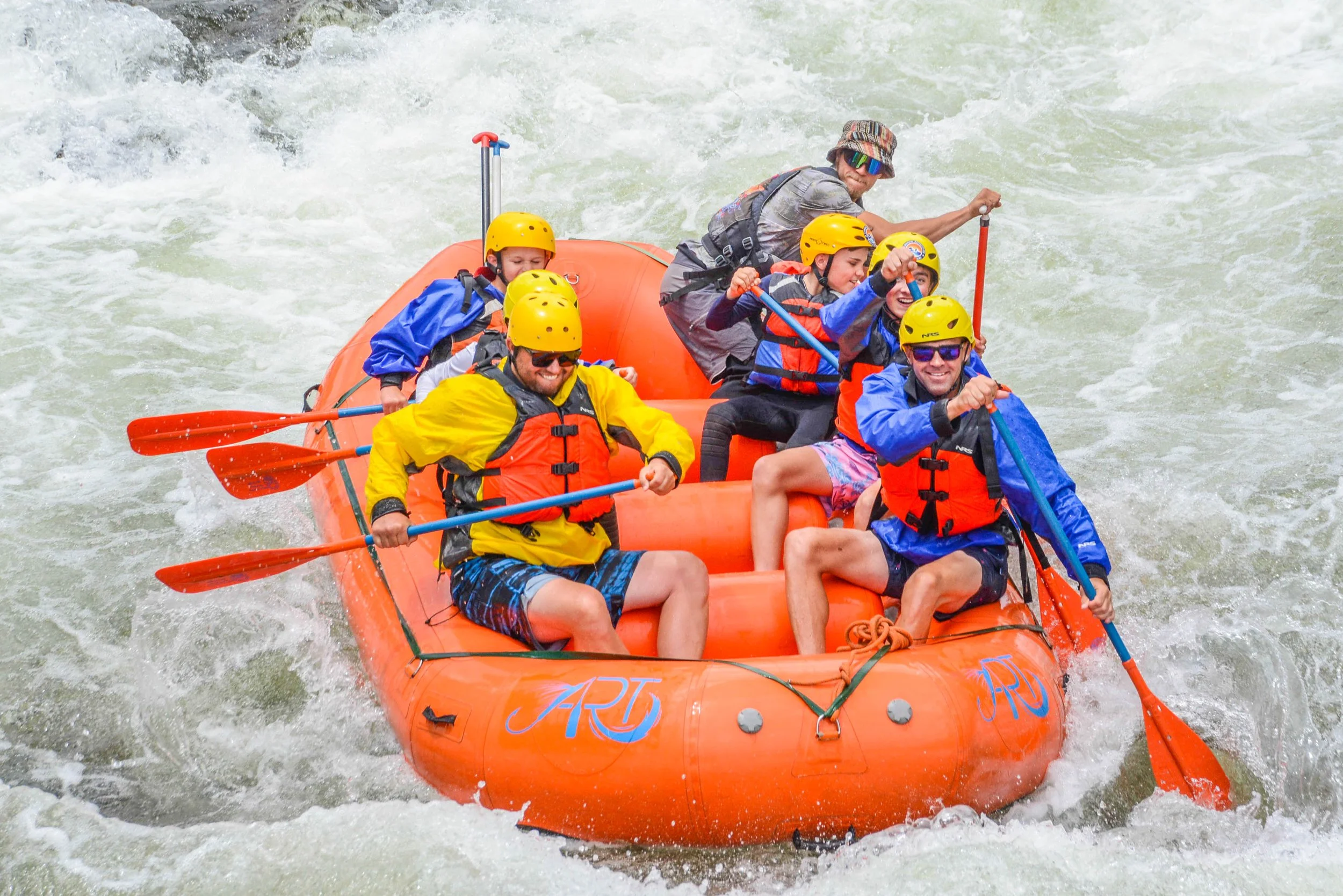 A group of six people white-water rafting on a river, wearing yellow helmets, life jackets, and colorful outdoor clothing, paddling with orange oars.