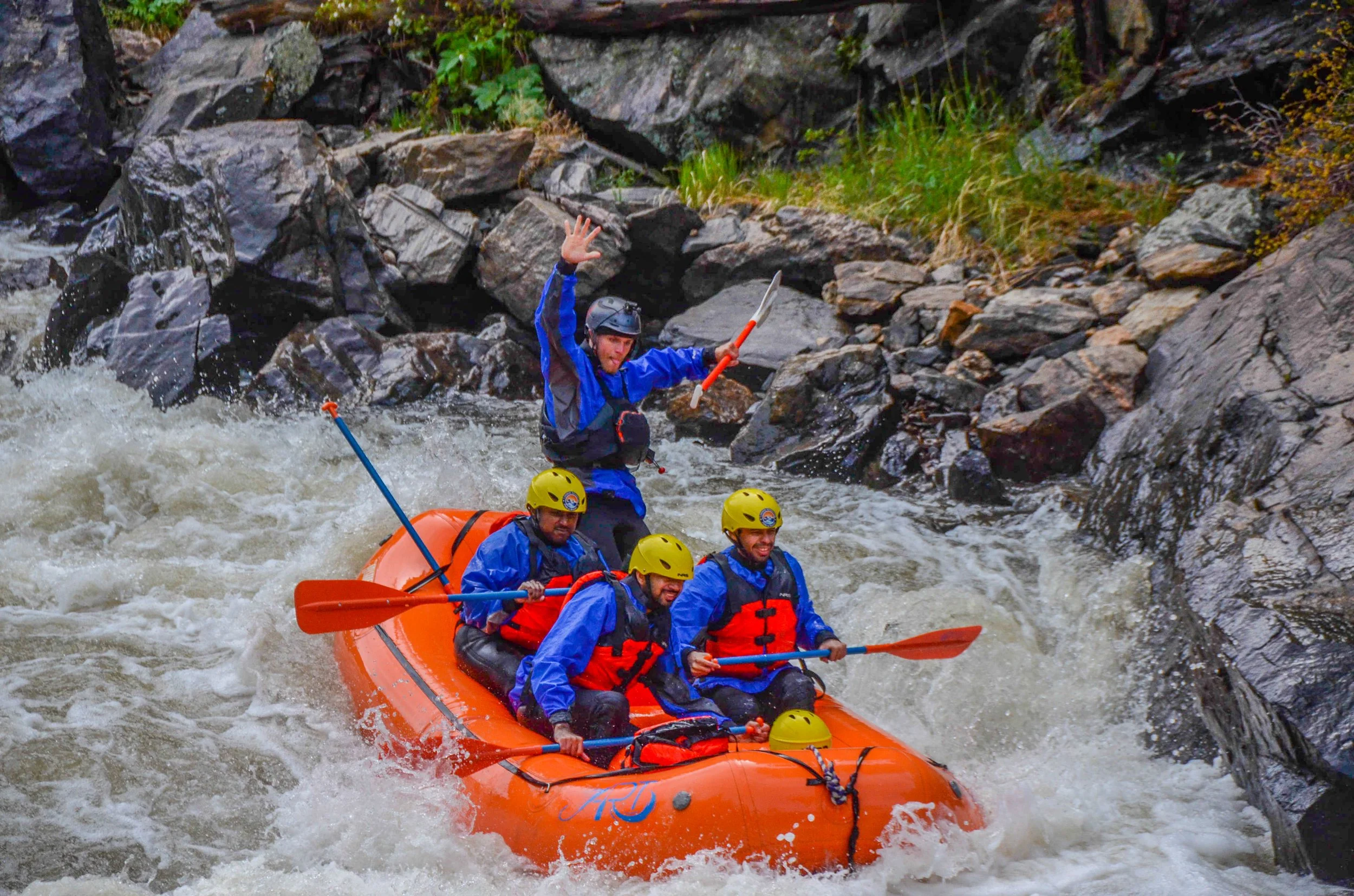 Four people in a bright orange raft navigating white water rapids on a river, wearing blue jackets, yellow helmets, and life vests, with one person standing and waving while holding an orange paddle, surrounded by rocks and turbulent water.