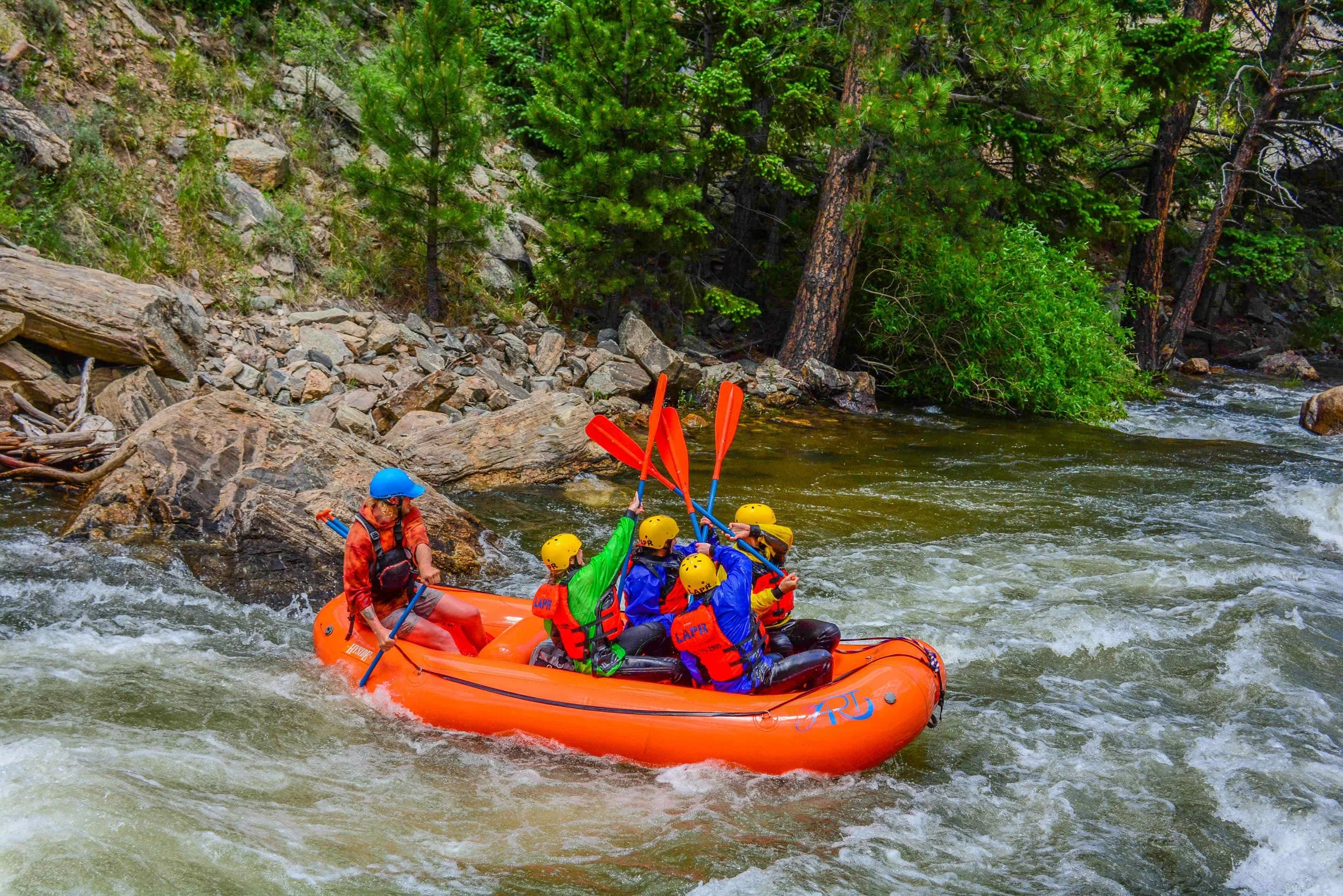 A group of six people in an orange inflatable raft with yellow helmets, paddling on a river with green trees and rocks along the shoreline.