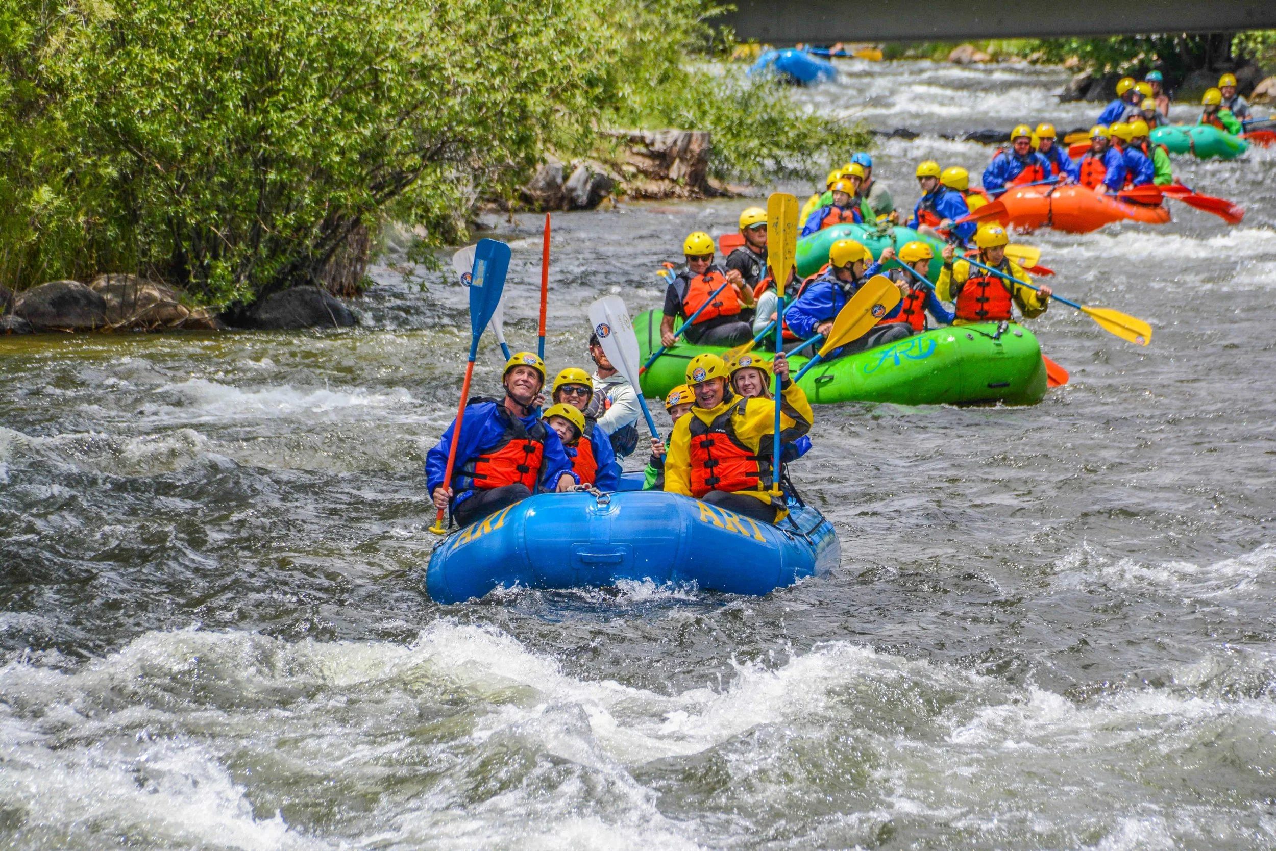 Group of people white water rafting on a river, wearing life jackets and yellow helmets, with some smiling and paddling.