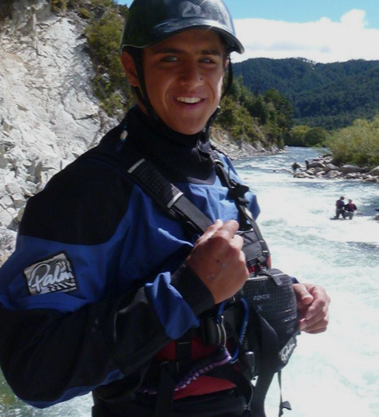 Young man smiling in outdoor river setting, wearing a black helmet and blue outdoor gear, holding a camera with a rugged landscape and other people in the background, expert guided rapid rafting tours at Idaho Springs Rafting Company, Colorado