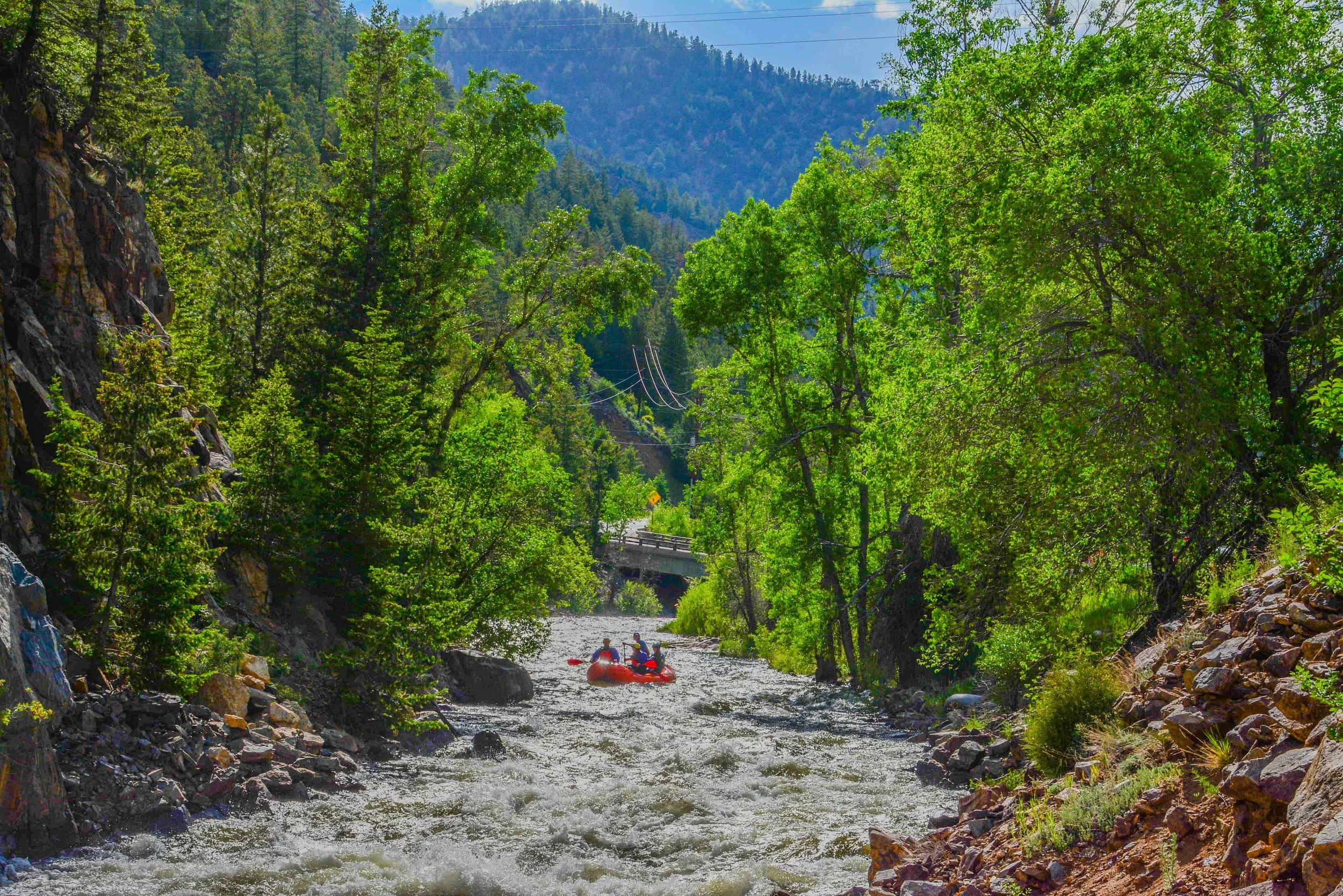 People in a red inflatable raft paddling down a rushing river surrounded by lush green trees and mountains in the background.