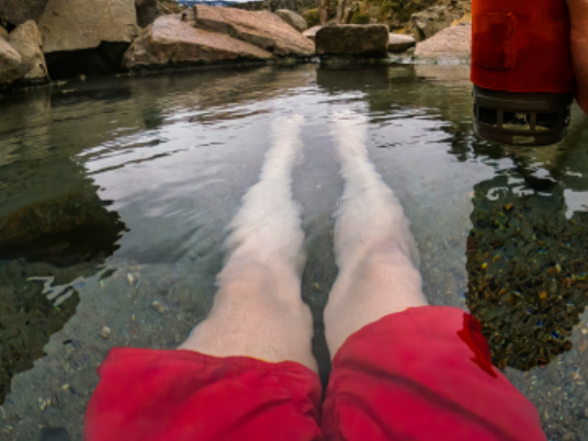 Person relaxing in a hot spring or pool with their legs submerged in water, rocks surrounding the area, and a red object nearby.