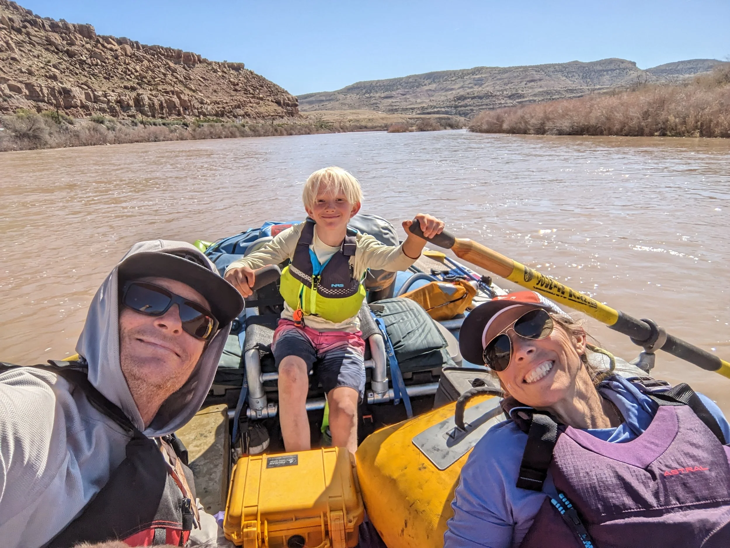 A family kayaking on a river in a desert landscape with rocky hills and sparse vegetation.