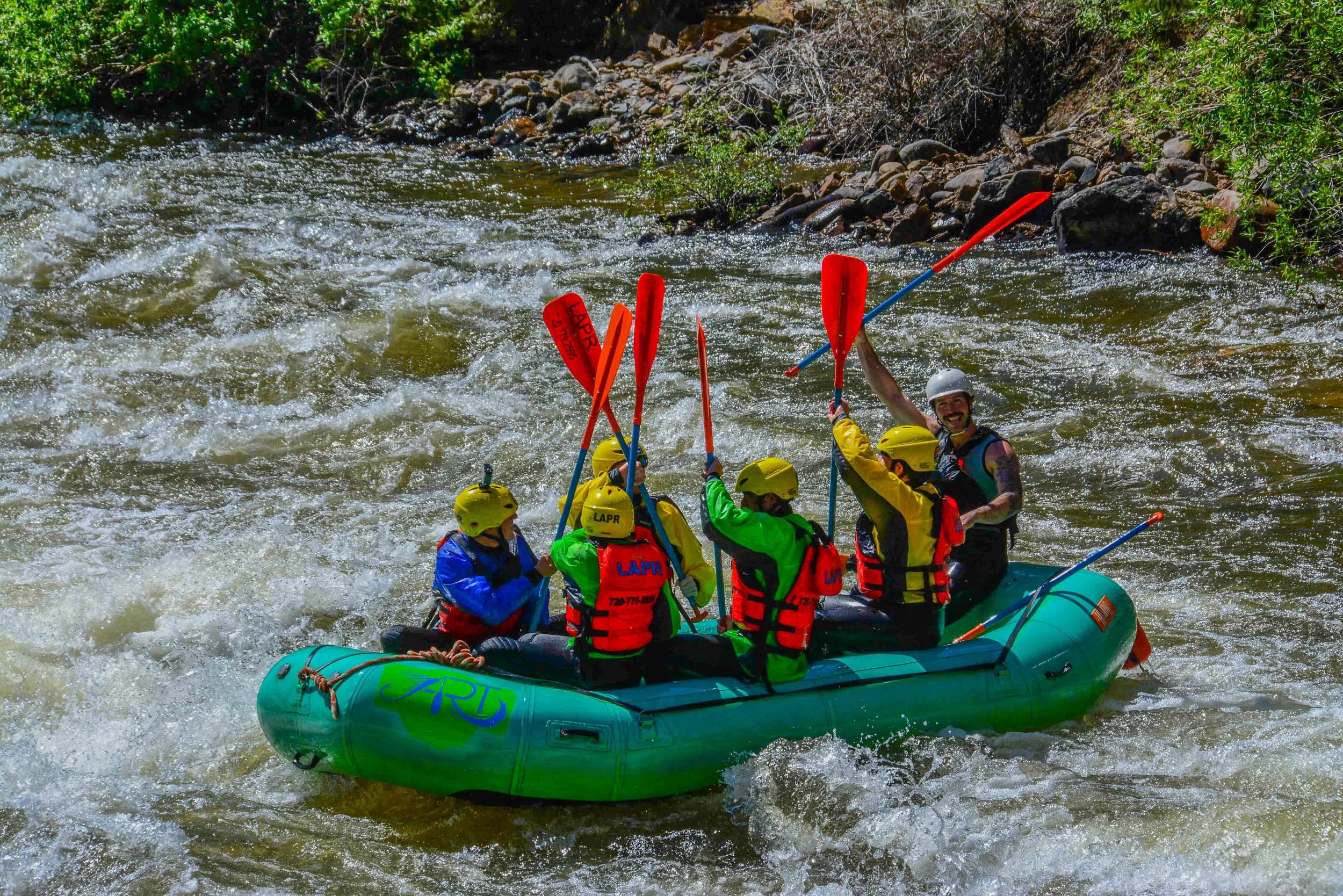 A group of people wearing helmets and life jackets white water rafting on a river with white rapids, surrounded by rocks and greenery.