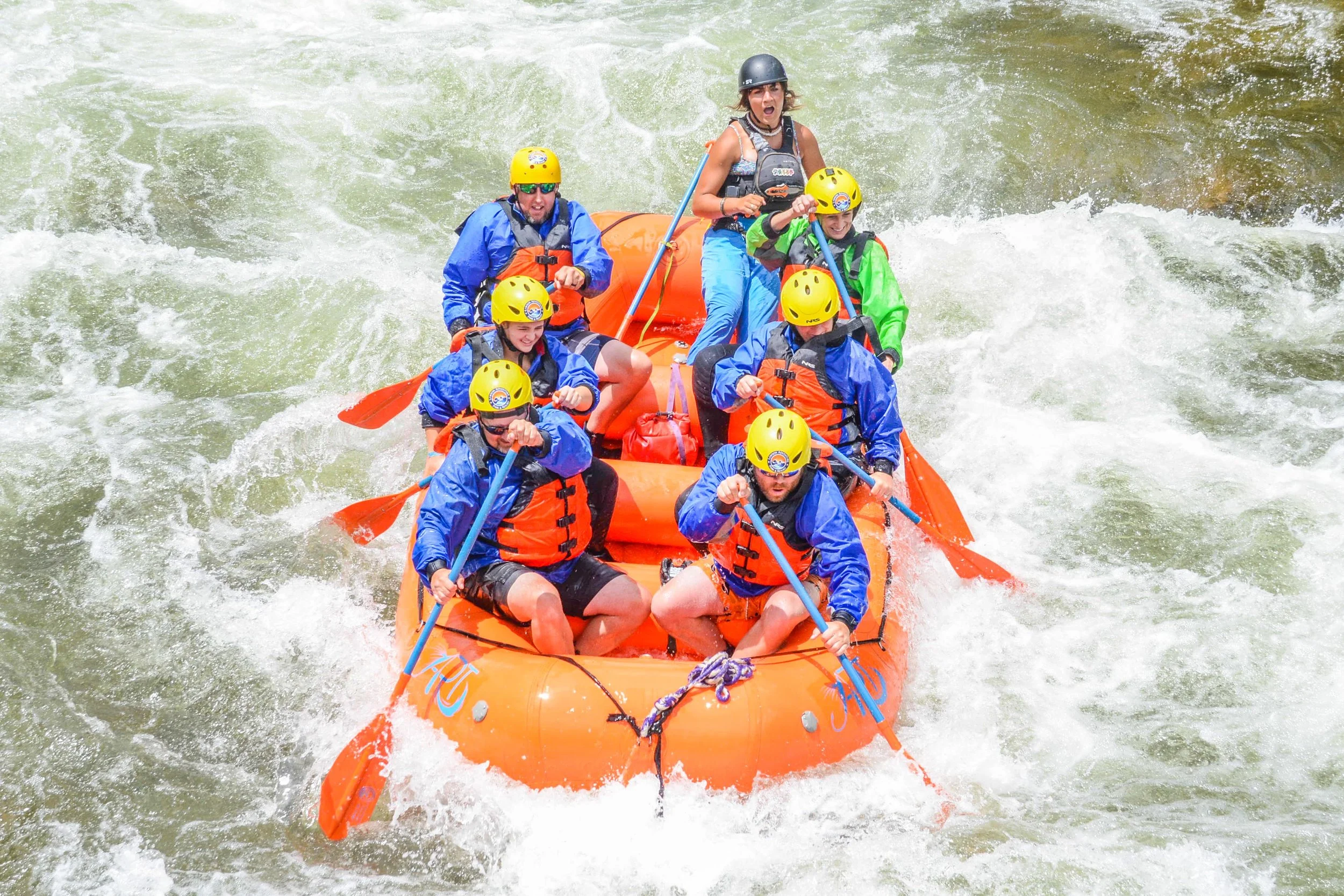 A group of seven people white water rafting on a river, wearing yellow helmets and life jackets, with one person standing and screaming, while others paddle through rough water.