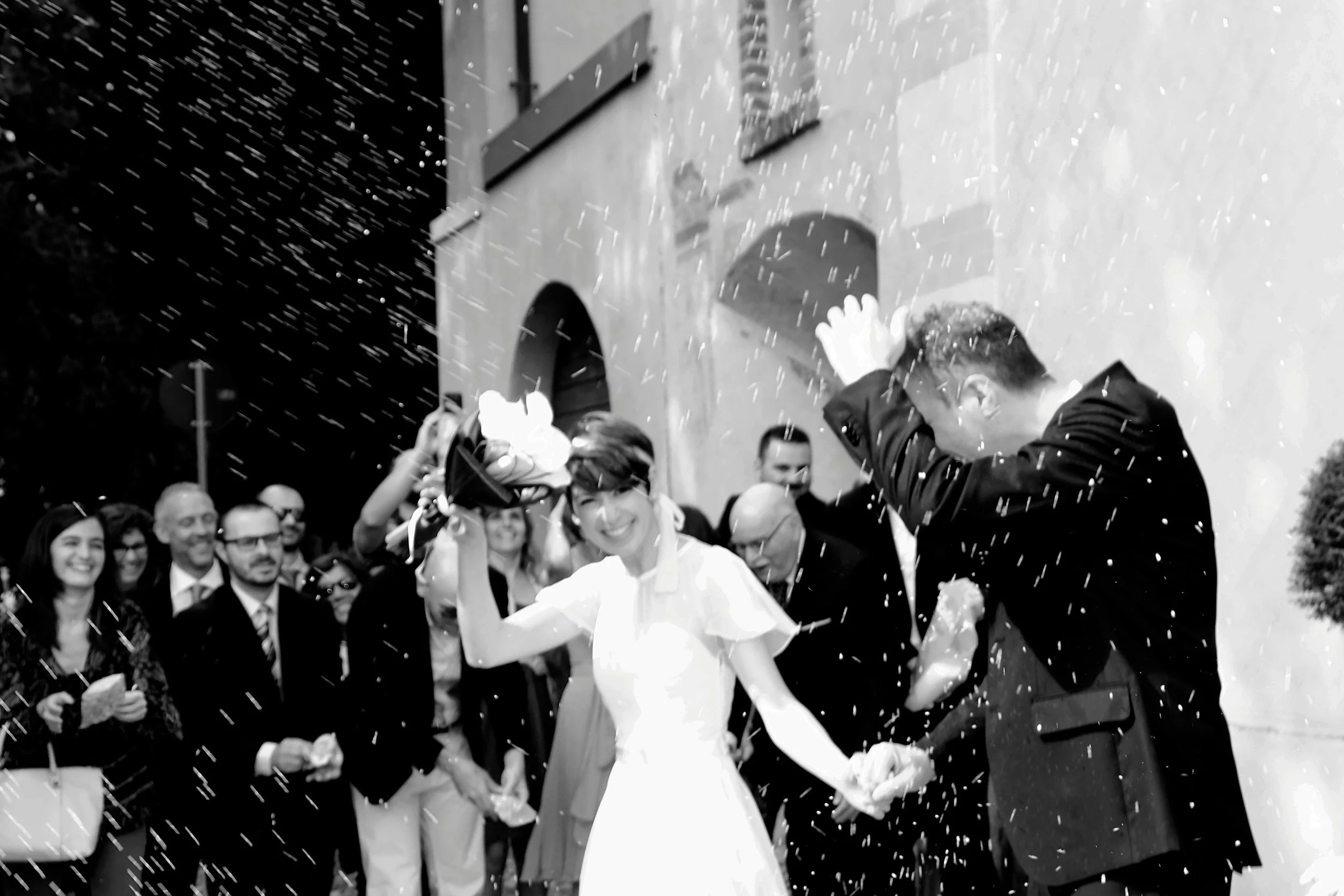 A bride and groom are celebrating outside a building, surrounded by smiling guests, as water or confetti is splashed over them.
