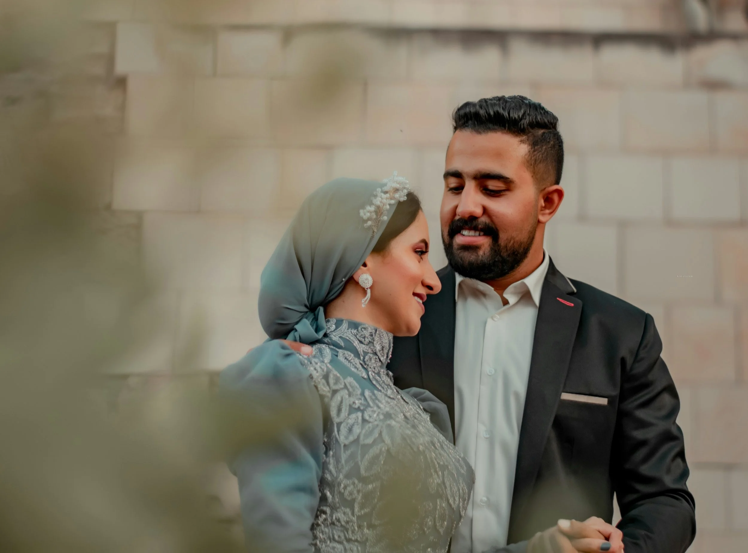 A couple dressed in formal attire sharing an intimate moment in front of a light-colored brick wall.