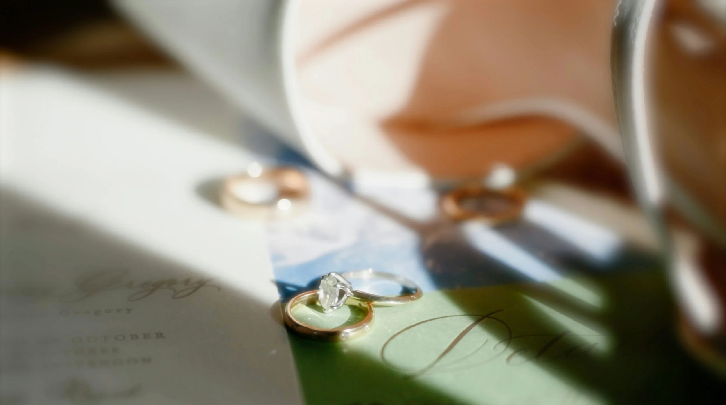 Close-up of a gold ring with a diamond, placed on a wedding invitation with multicolored shadows cast by jewelry and paper.