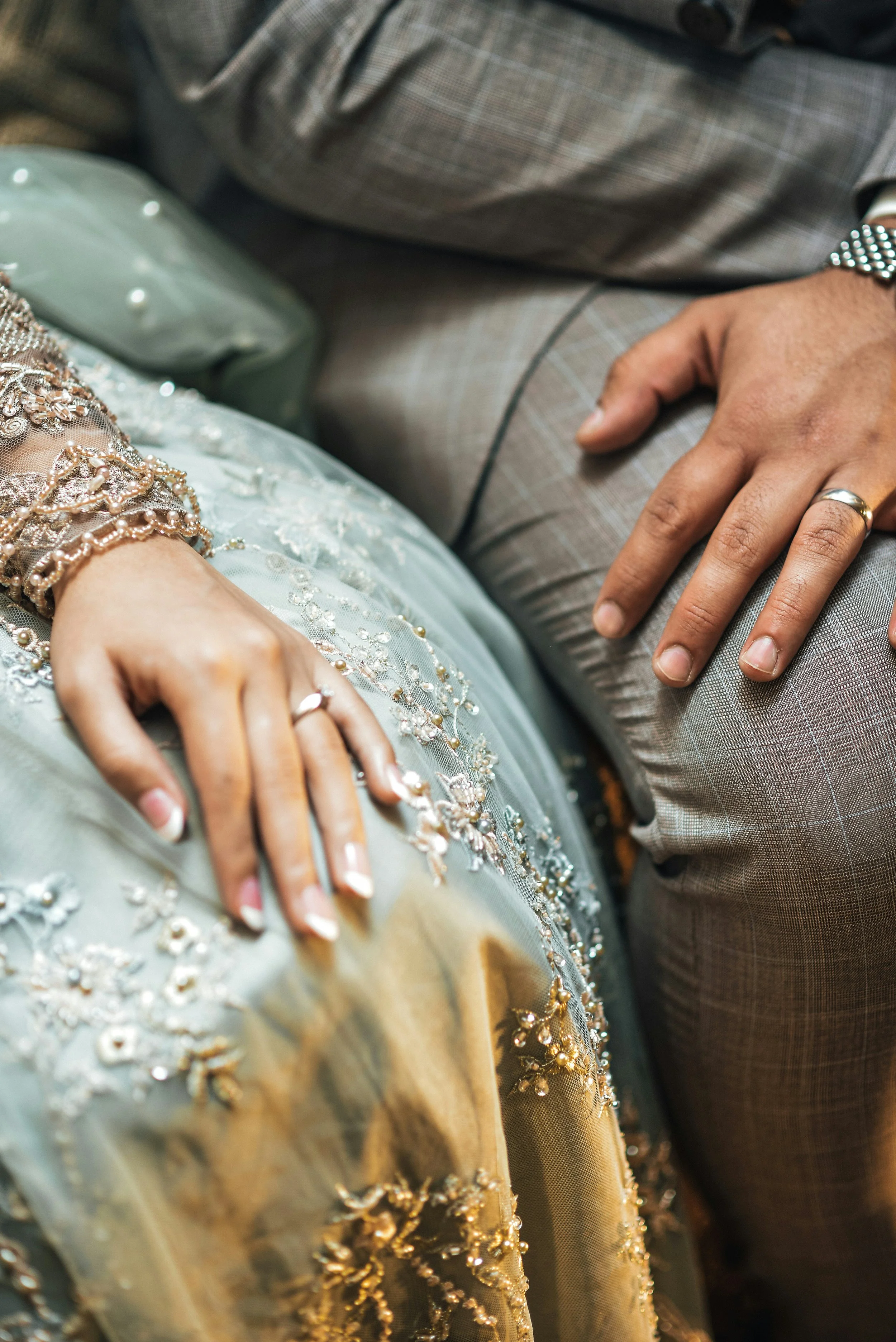 Close-up of a bride and groom's hands, showing wedding rings, seated together. The bride wears a dress with intricate embroidery and beading, and the groom wears a gray plaid suit.