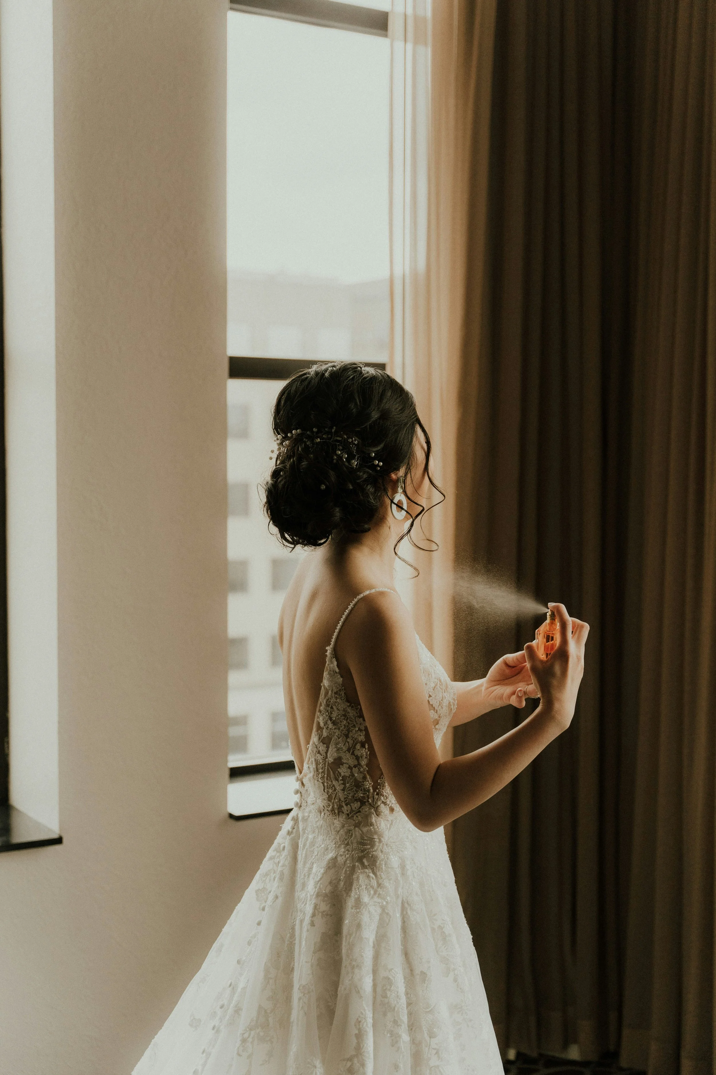 Bride in wedding dress spraying perfume in a bright hotel room near a window with beige curtains.