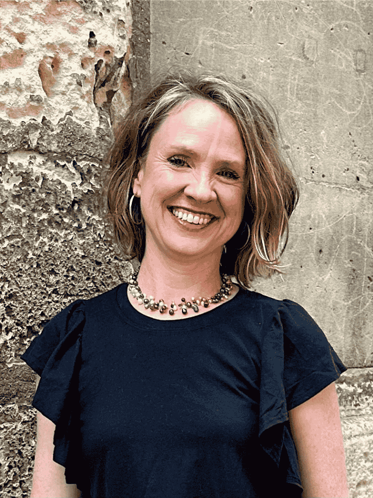 Carrie Henry with shoulder-length wavy hair, wearing hoop earrings, a beaded necklace, and a black top with ruffle details on the sleeves, smiling while standing outdoors near a textured stone wall.