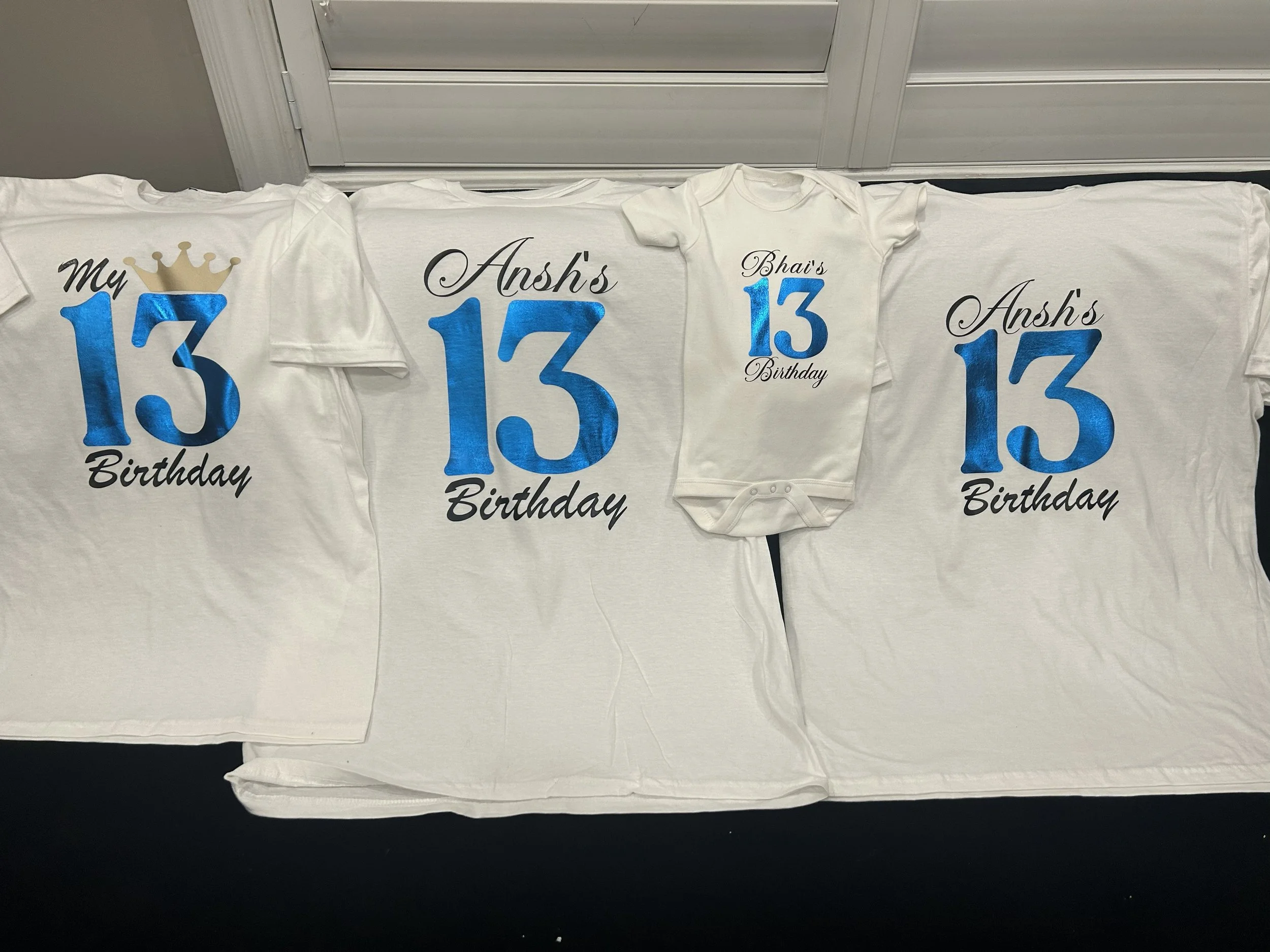 White T-shirts and a baby onesie with blue numbers and black cursive text for a birthday celebration, displayed on a white sheet.