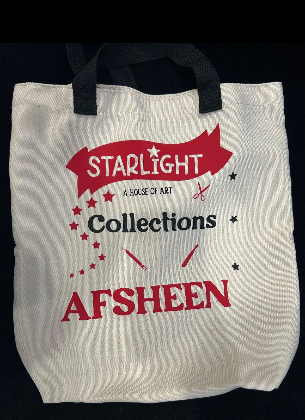 A white tote bag with black handles displaying a red and black design that includes text about art collections, scissors, and a needle.