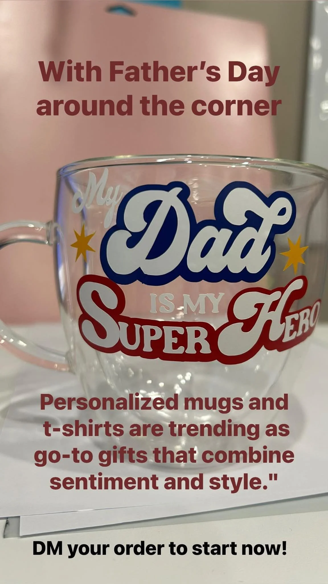 A clear glass mug with colorful text that reads "My Dad is My Superhero". In the background, there is a pink sign with text about personalized mugs and T-shirts for Father's Day.