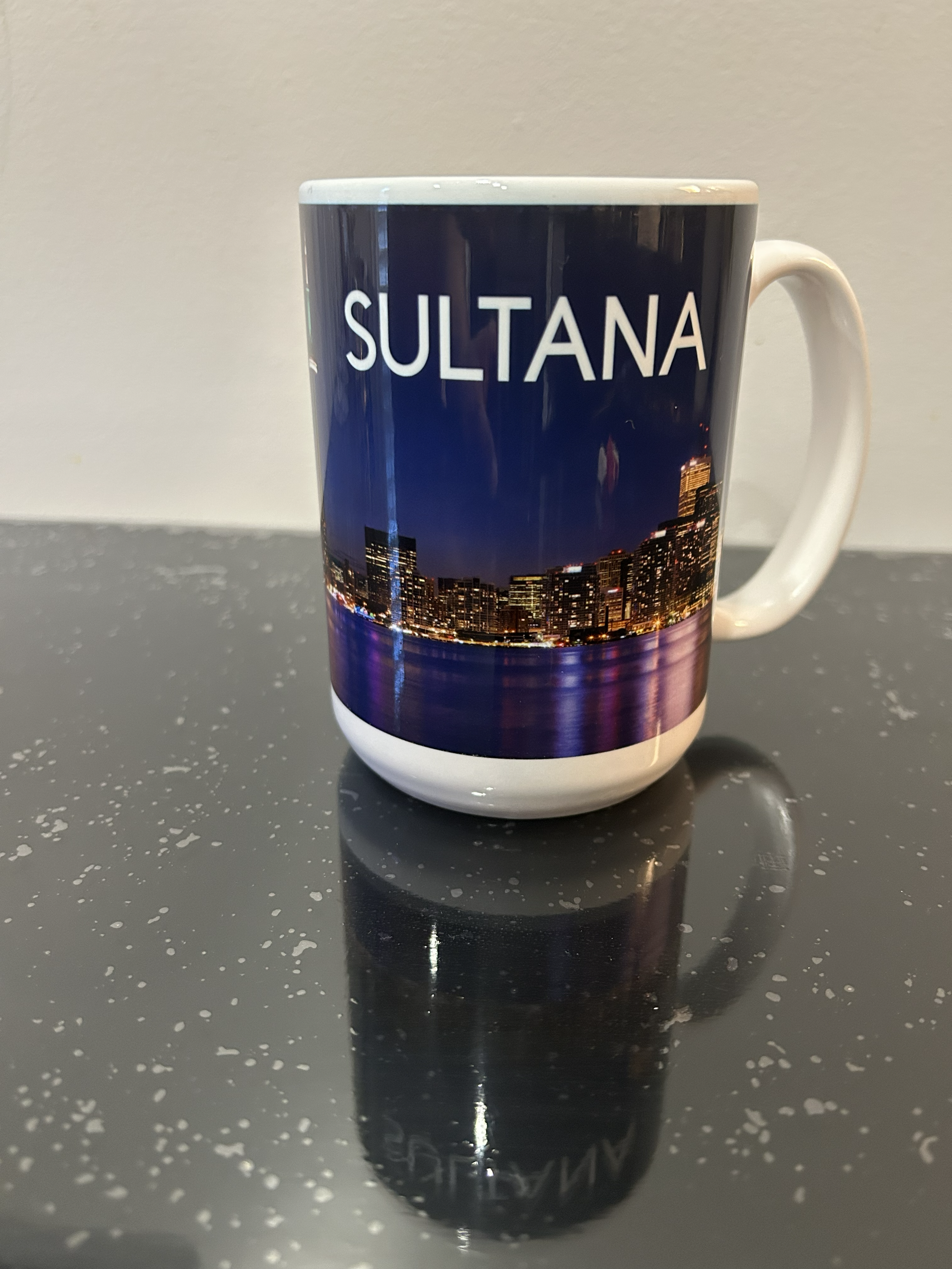 A coffee mug with a cityscape and night sky design, with the word "SULTANA" printed on it, placed on a dark speckled surface.