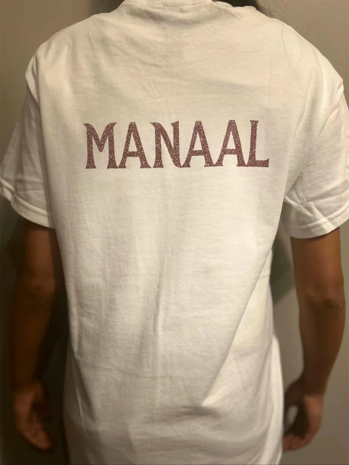 Person wearing a white T-shirt with the word "MANAAAL" written in sparkly letters on the back.