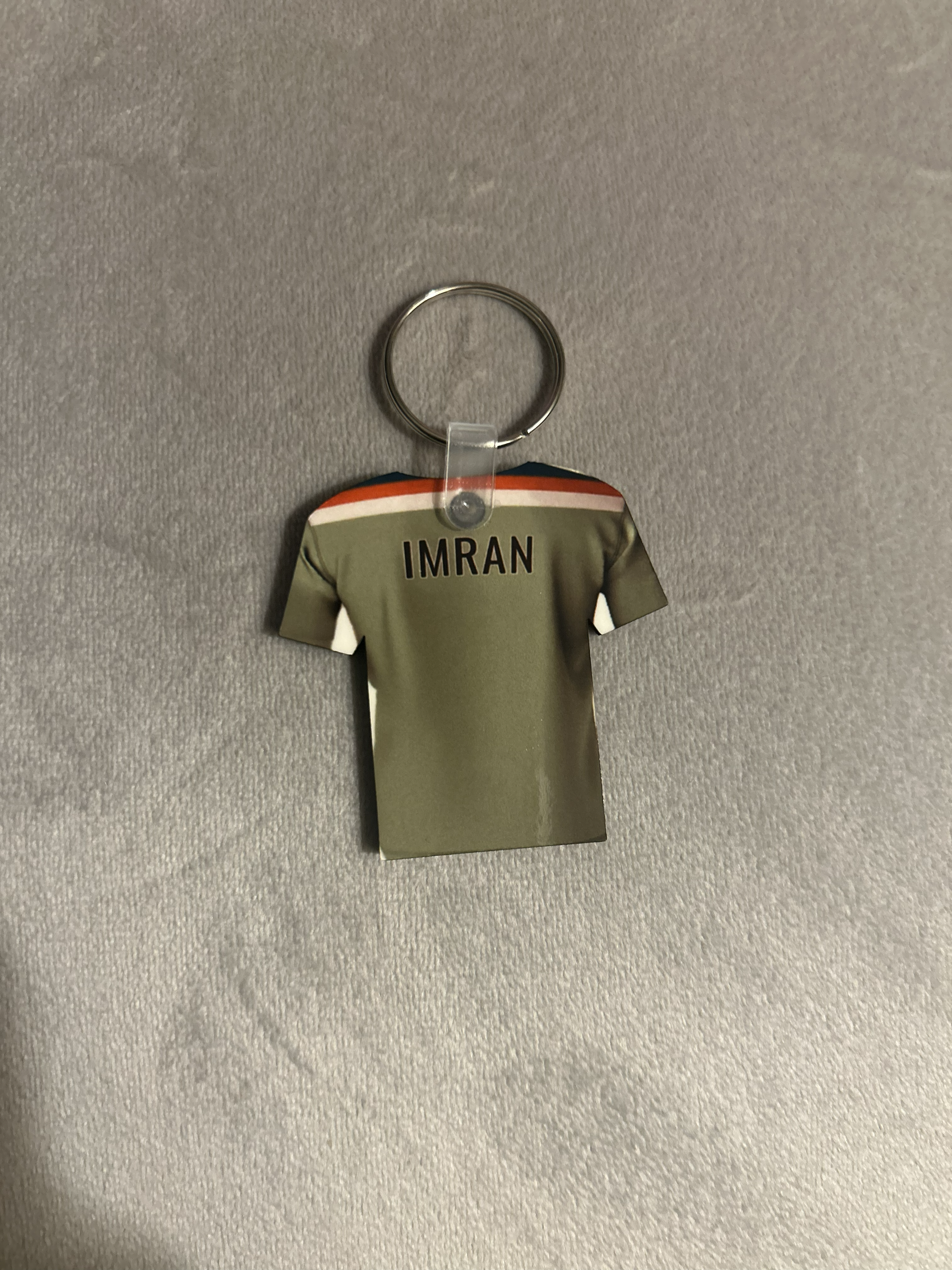 Keychain with a paper cutout of a uniformed figure and the name 'IMRAN' attached to a textured grey surface.
