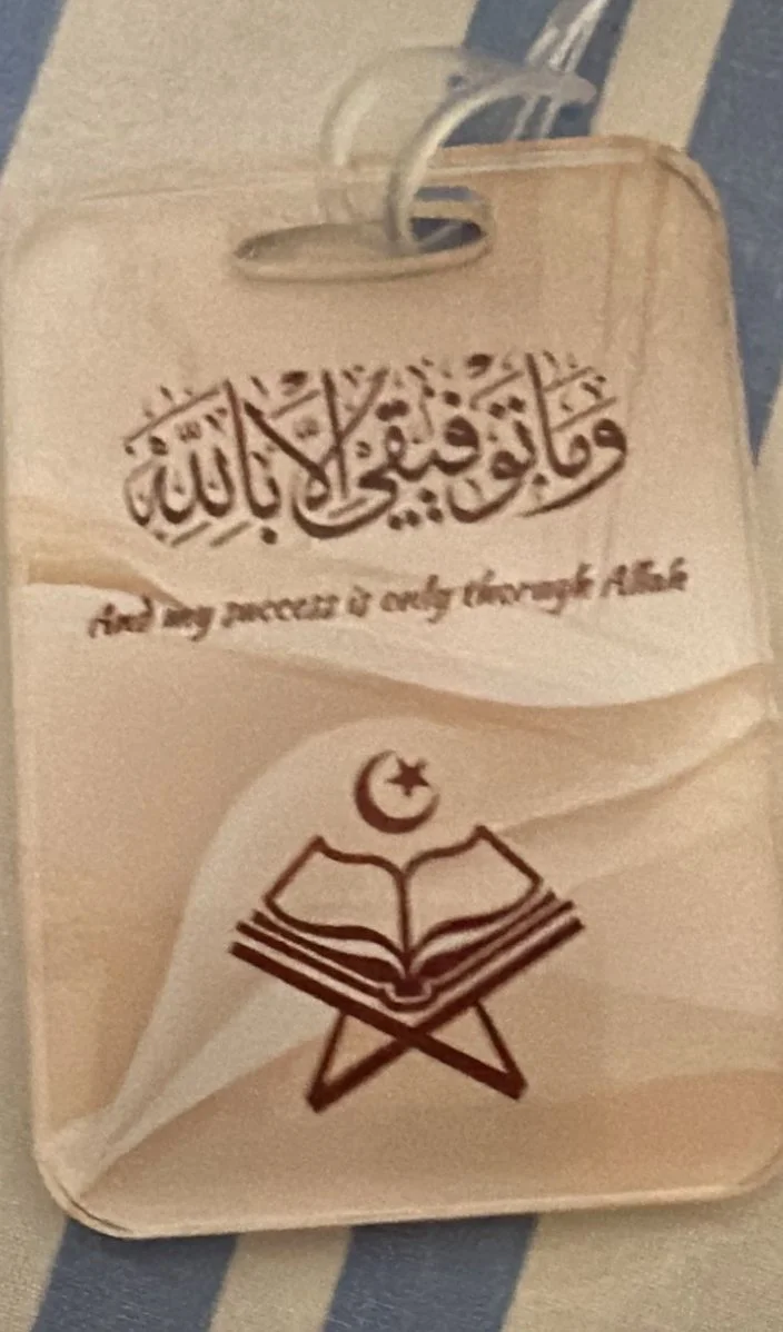 A beige notebook with Arabic calligraphy and a phrase in English reading 'and my success is only through Allah.' It also has a drawing of an open book with a crescent moon and star above it. The notebook is on a blue and white striped fabric and has 