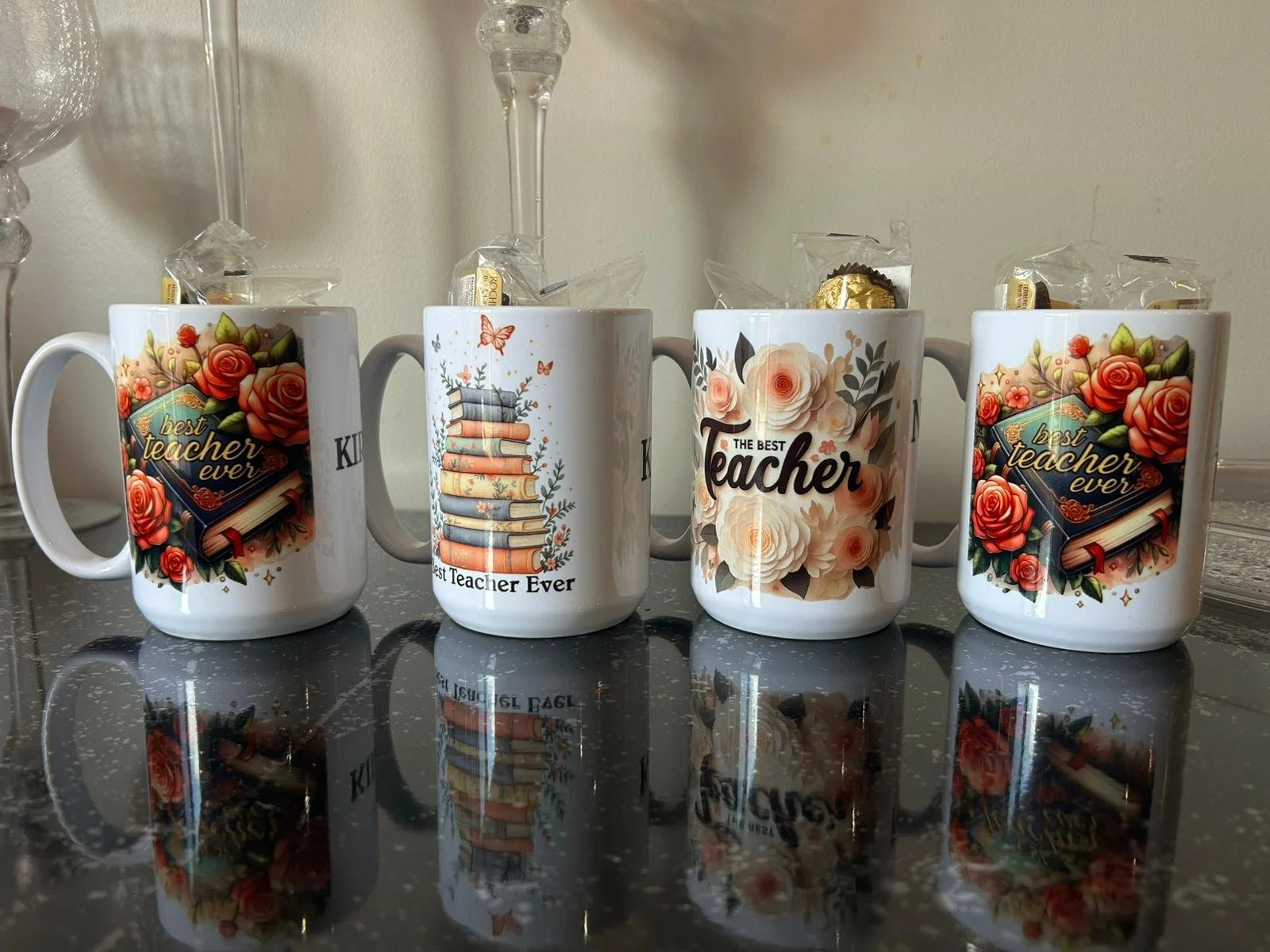 Four coffee mugs on a black speckled surface with gold reflections, each decorated with floral or book-themed designs and phrases about teachers.