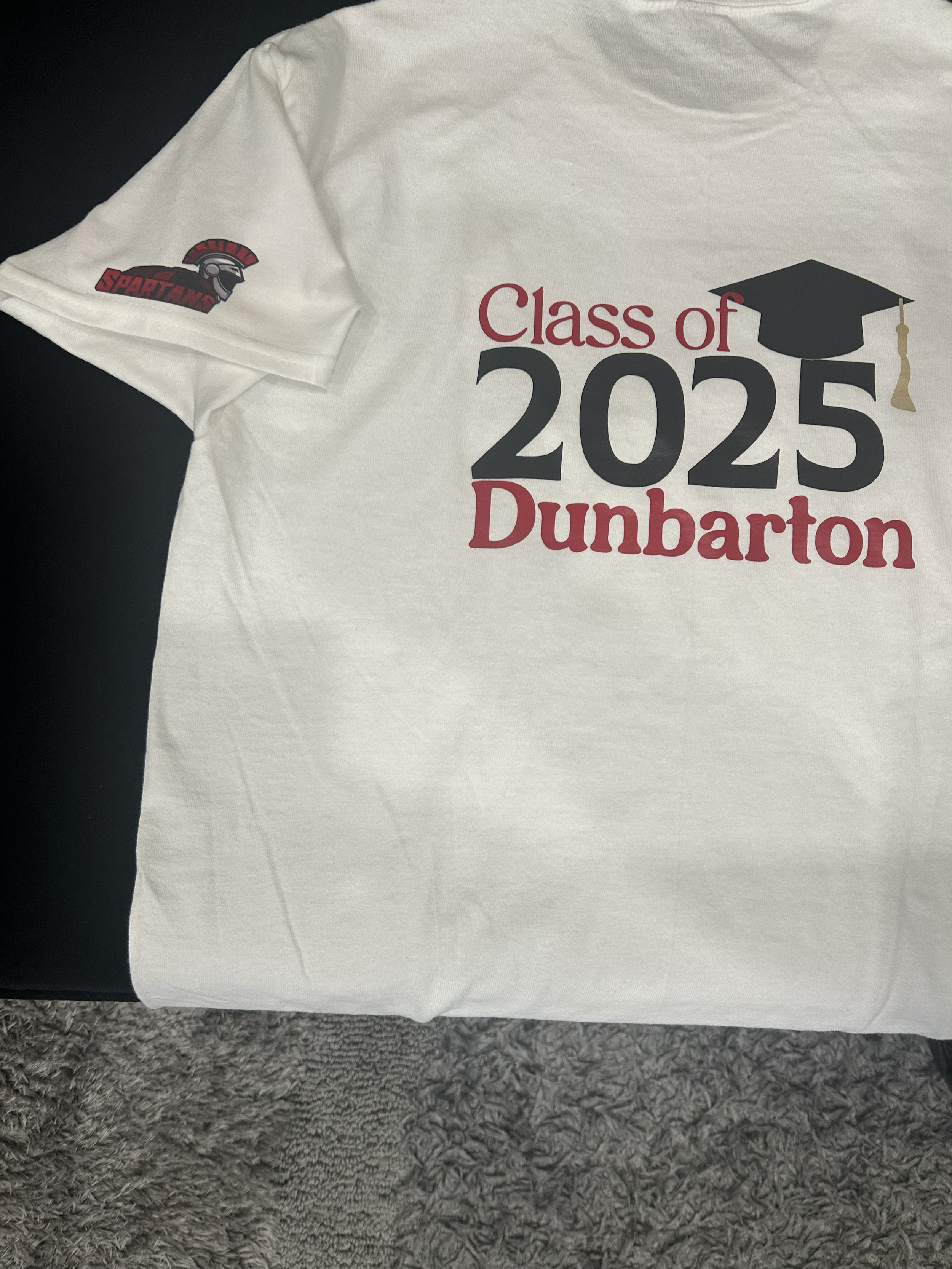 A white T-shirt with the words "Class of 2025 Dunbarton" printed on it, with a black graduation cap icon and a beige tassel. The T-shirt also has a Spartan helmet logo with the word "SPARTANS" on the sleeve.