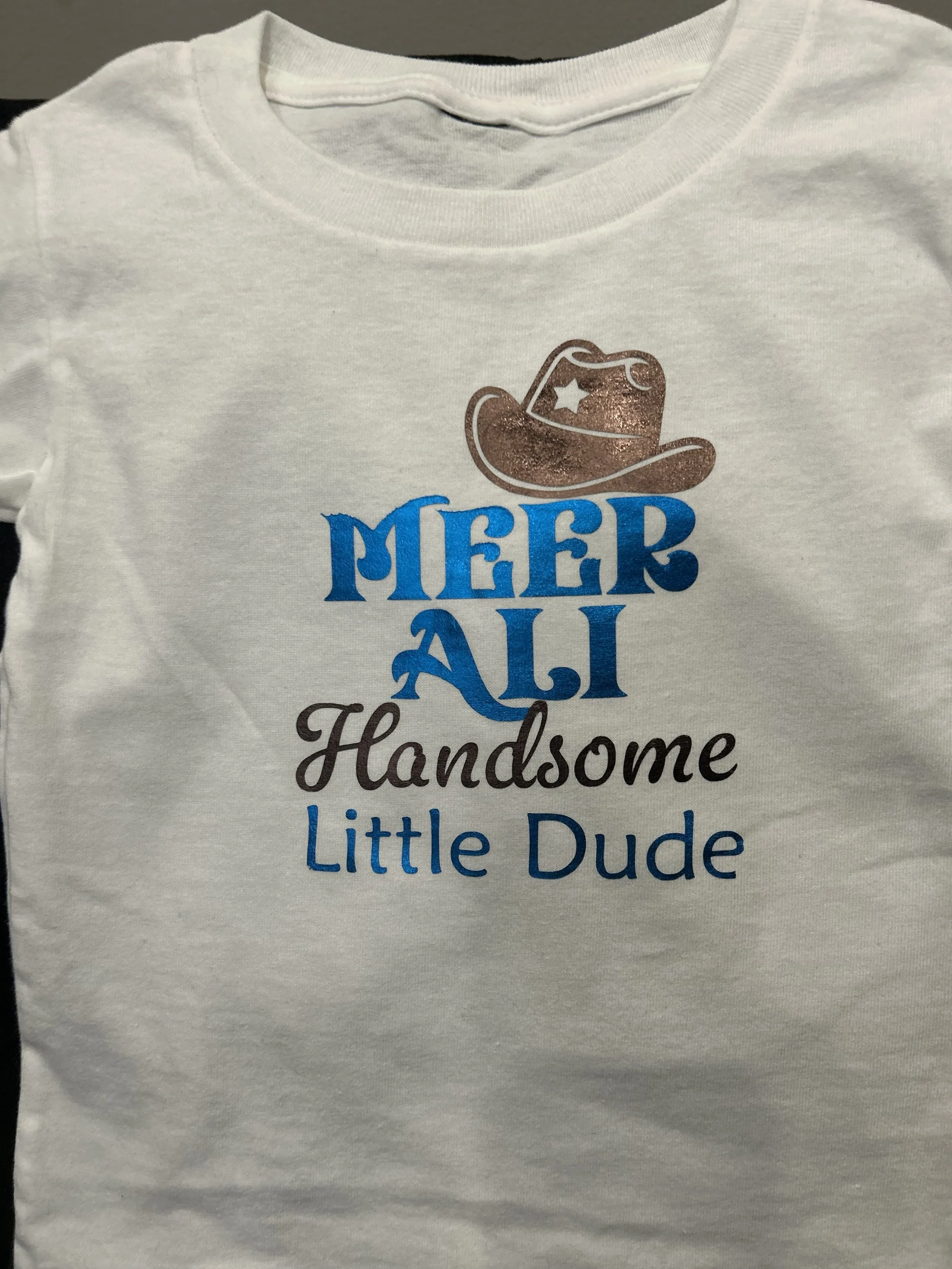 White T-shirt with a graphic of a cowboy hat and star, and text that reads 'Merry All Handsome Little Dude'.