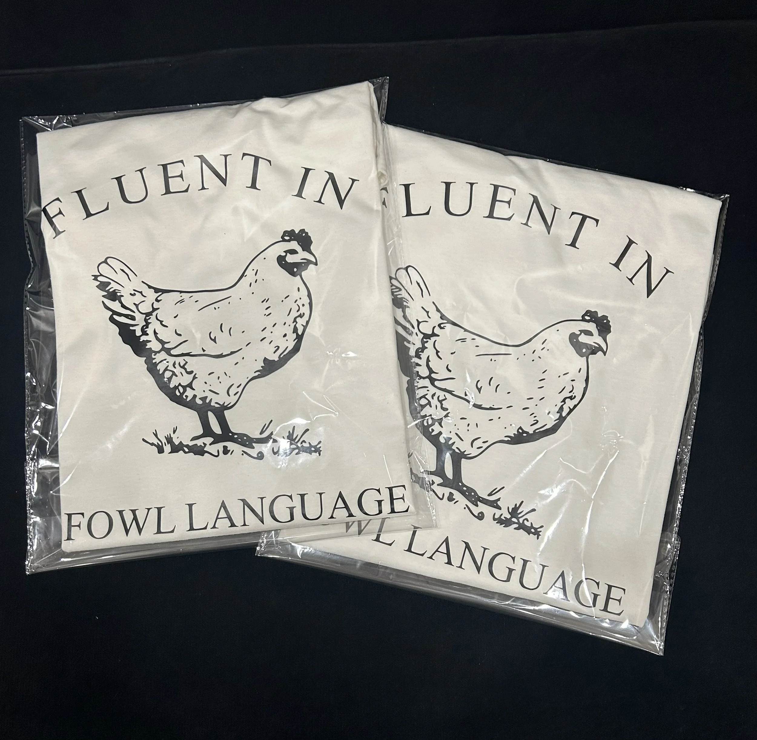 Close-up of a fabric flag with a black outline illustration of a chicken and the text 'FLUENT IN FOWL LANGUAGE' in black, packaged in a clear plastic sleeve.