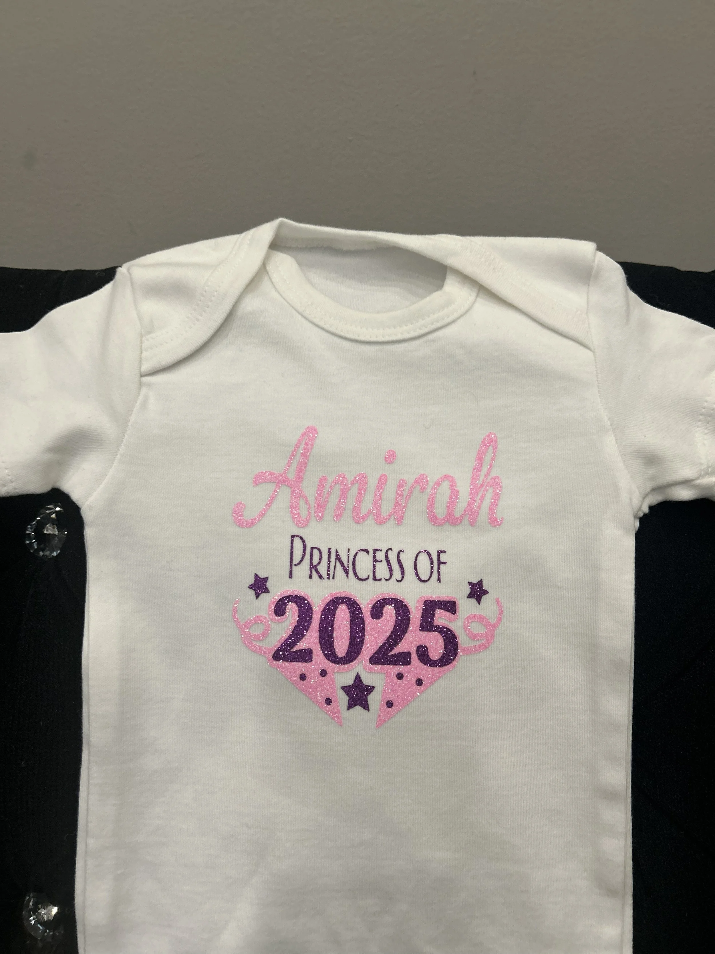 A white T-shirt with pink and purple text reading "Honest" and "Princess of 2025" decorated with purple and pink stars, laid on a black surface with sequin-like embellishments.