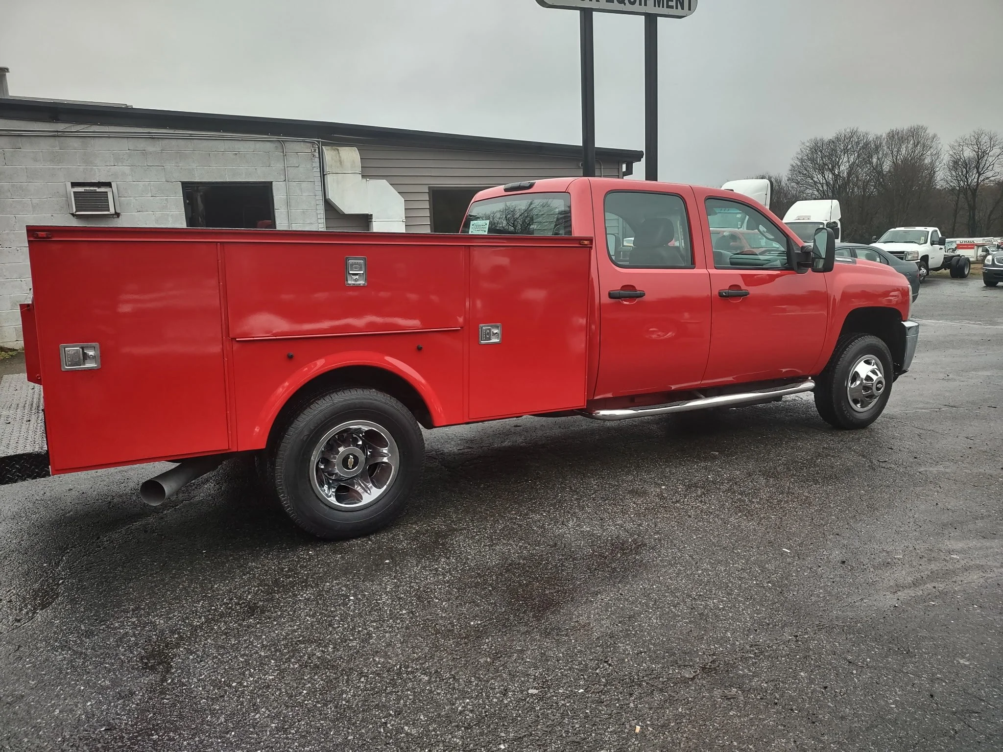 Red utility truck with multiple storage compartments parked on asphalt, with other vehicles and a gray building in the background. Body paint job for heavy trucks.