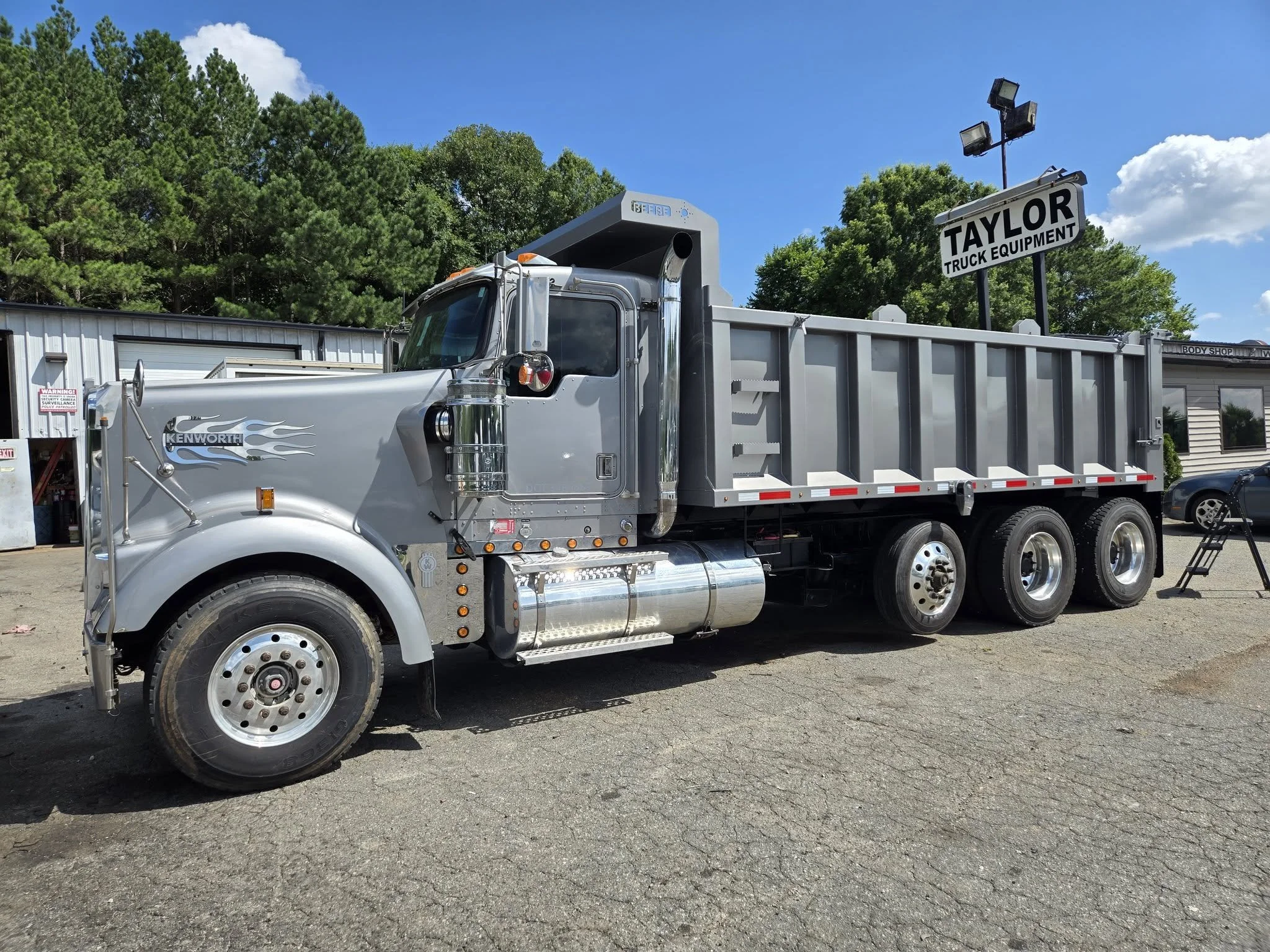 Silver Kenworth dump truck parked outside a building with a Taylor Truck Equipment sign, trees, and a blue sky in the background. Body paint job for a used truck company.
